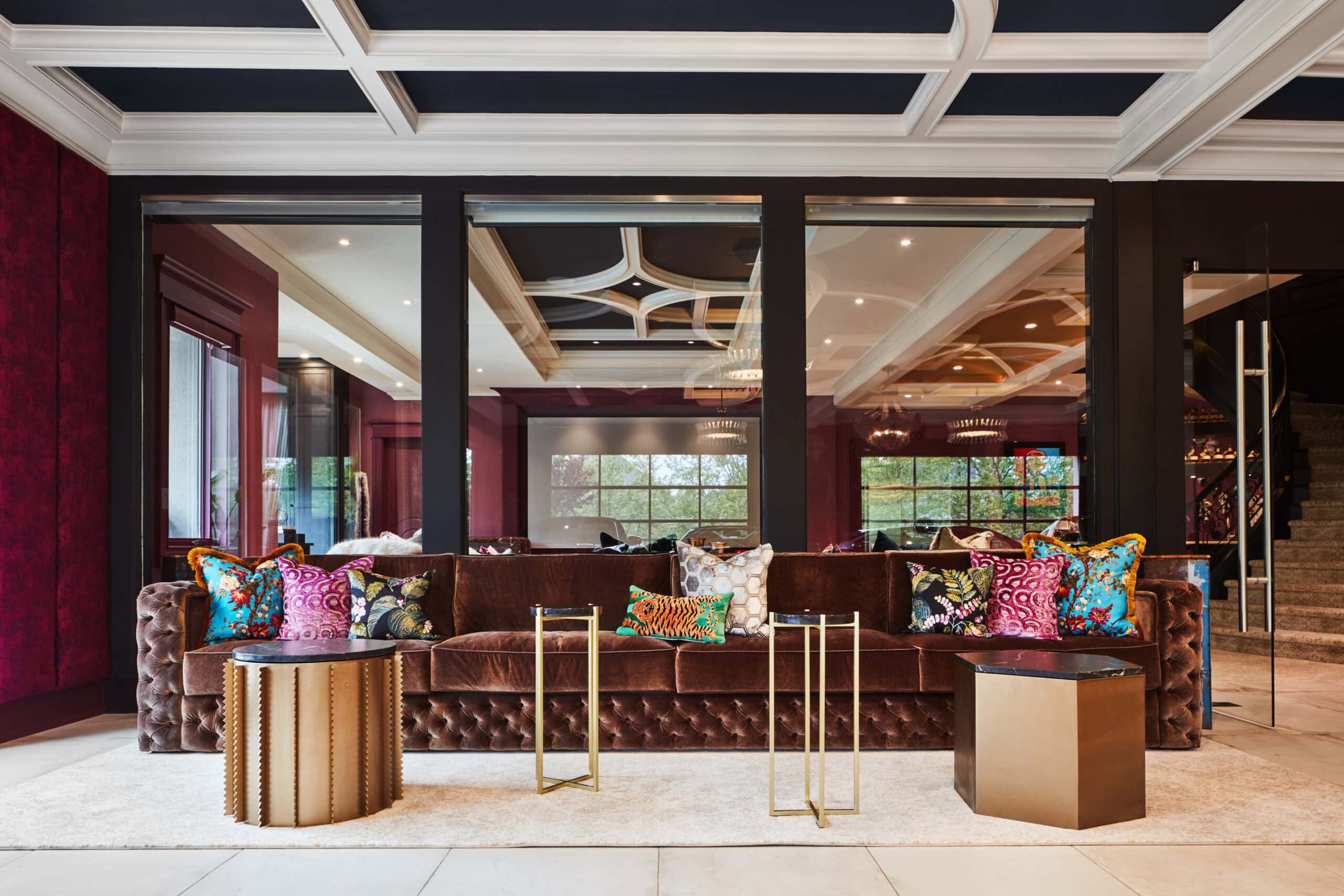 A modern living room with a brown tufted sofa, colorful patterned pillows, geometric side tables, and a coffered ceiling reflected in large glass panels.