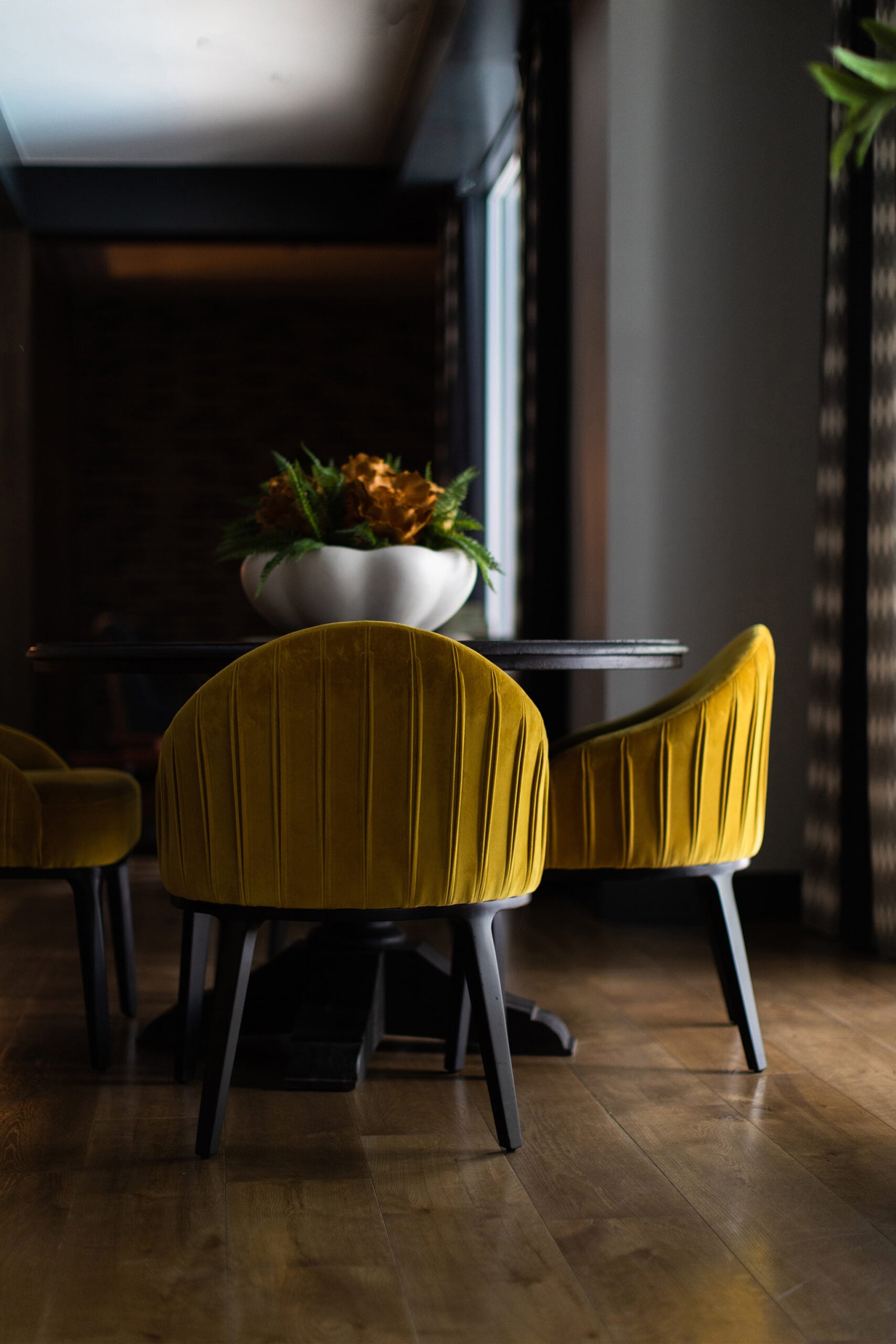 A dark dining area by Crimson Design Group features a round table with a white bowl of plants as the centerpiece and yellow velvet chairs on a wooden floor.
