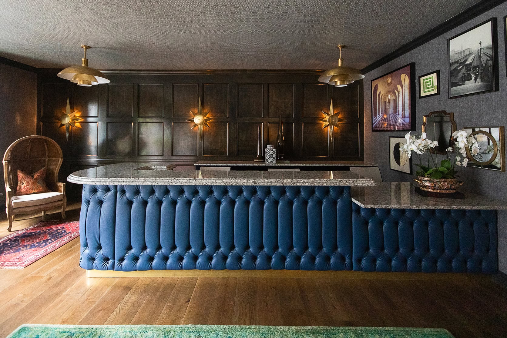 A bar with a tufted blue front, granite countertop, wood-paneled wall, two gold pendant lights, framed artwork, and decorative plants on a wooden floor.