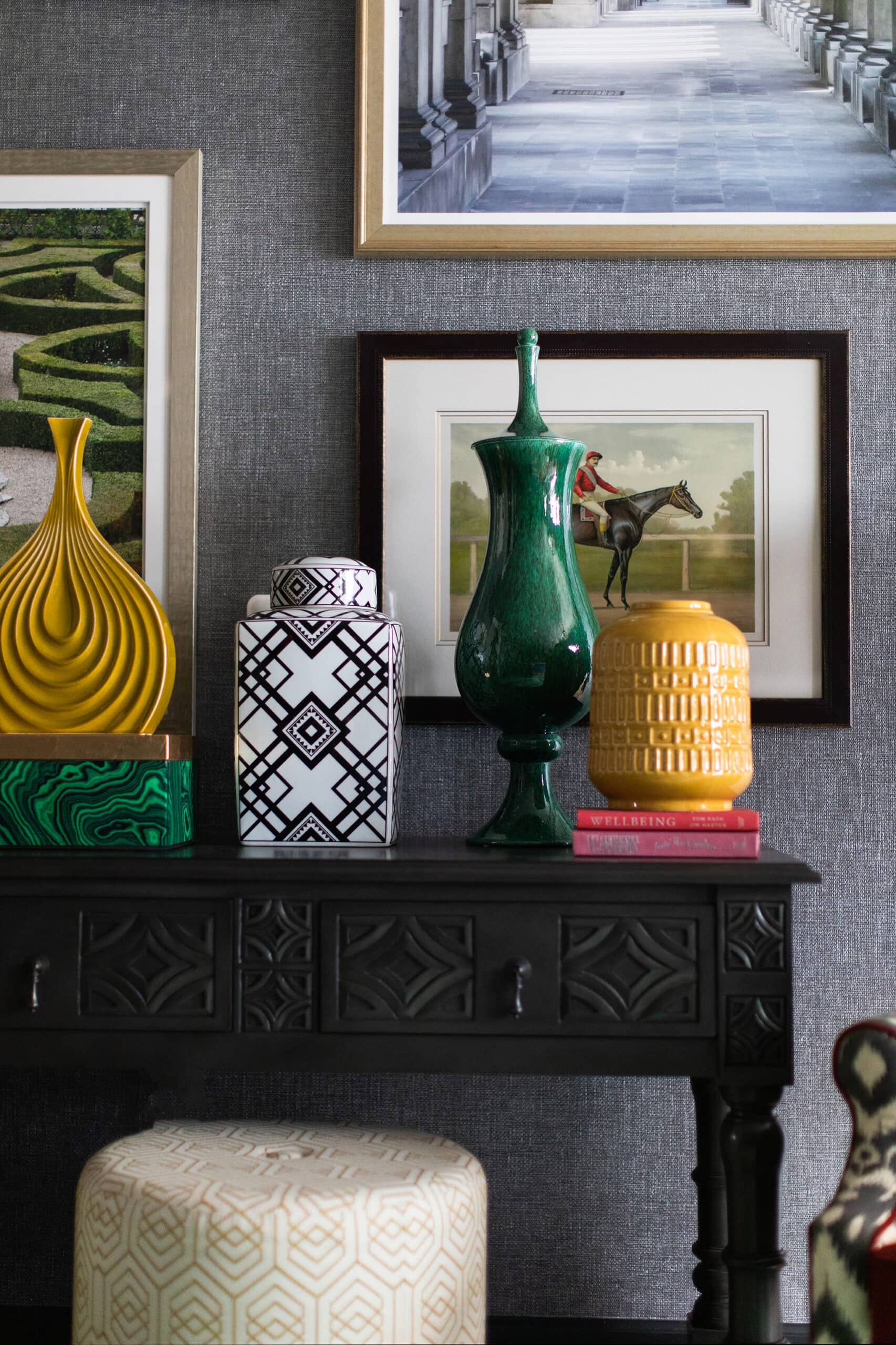 A black table with patterned vases and books is set against a gray wall with framed artwork and photographs.
