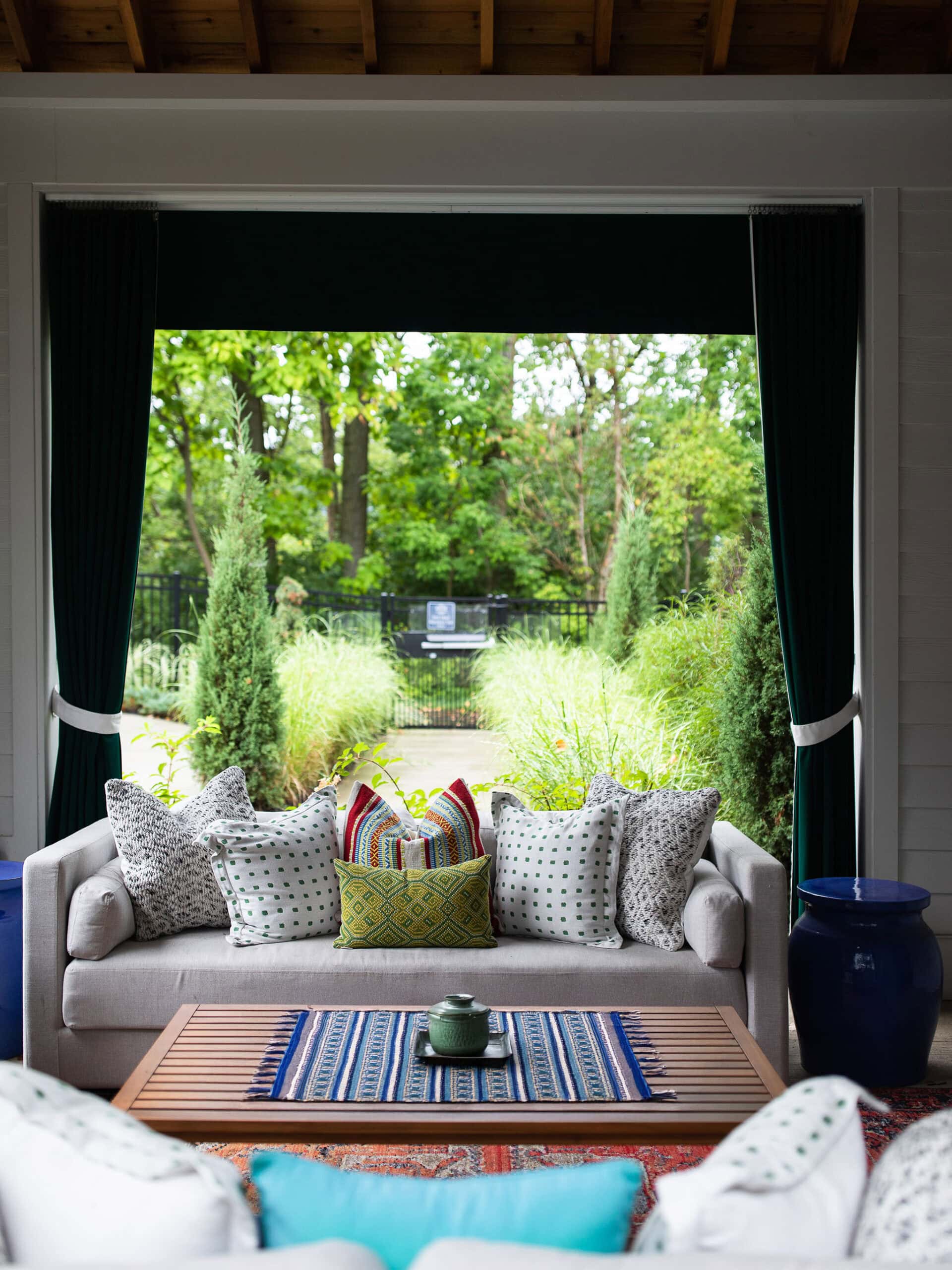 A beige sofa with patterned cushions sits on a covered patio, facing a wooden coffee table, with green plants and trees visible outside.