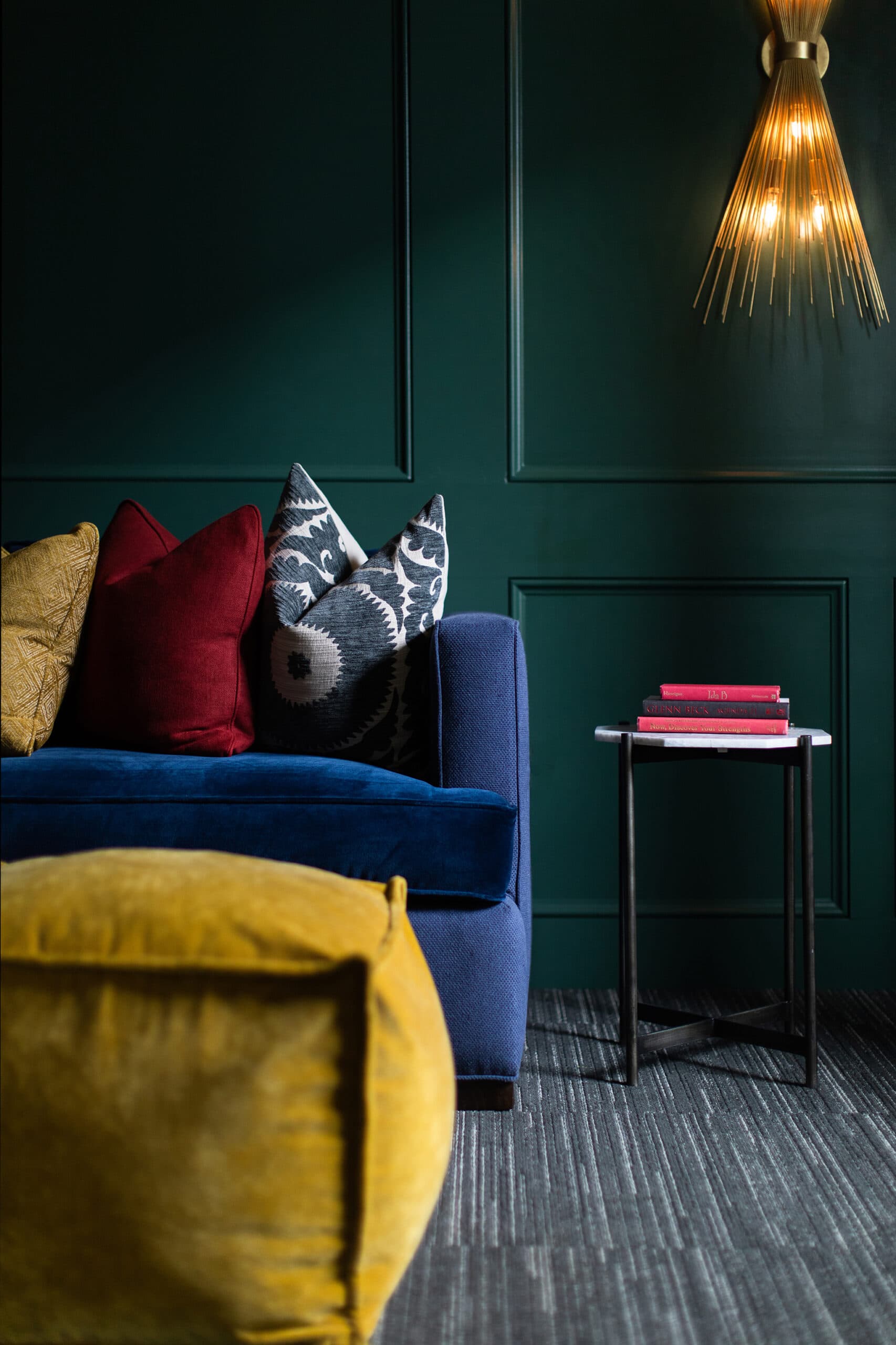 A modern living room with a dark green wall, blue sofa with colorful cushions, yellow ottoman, small side table with books, and a gold wall sconce.