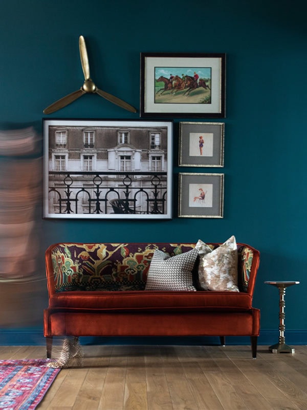A red velvet sofa with patterned cushions sits against a teal wall, which is decorated with framed art and a brass propeller above. A small metal side table is nearby.
