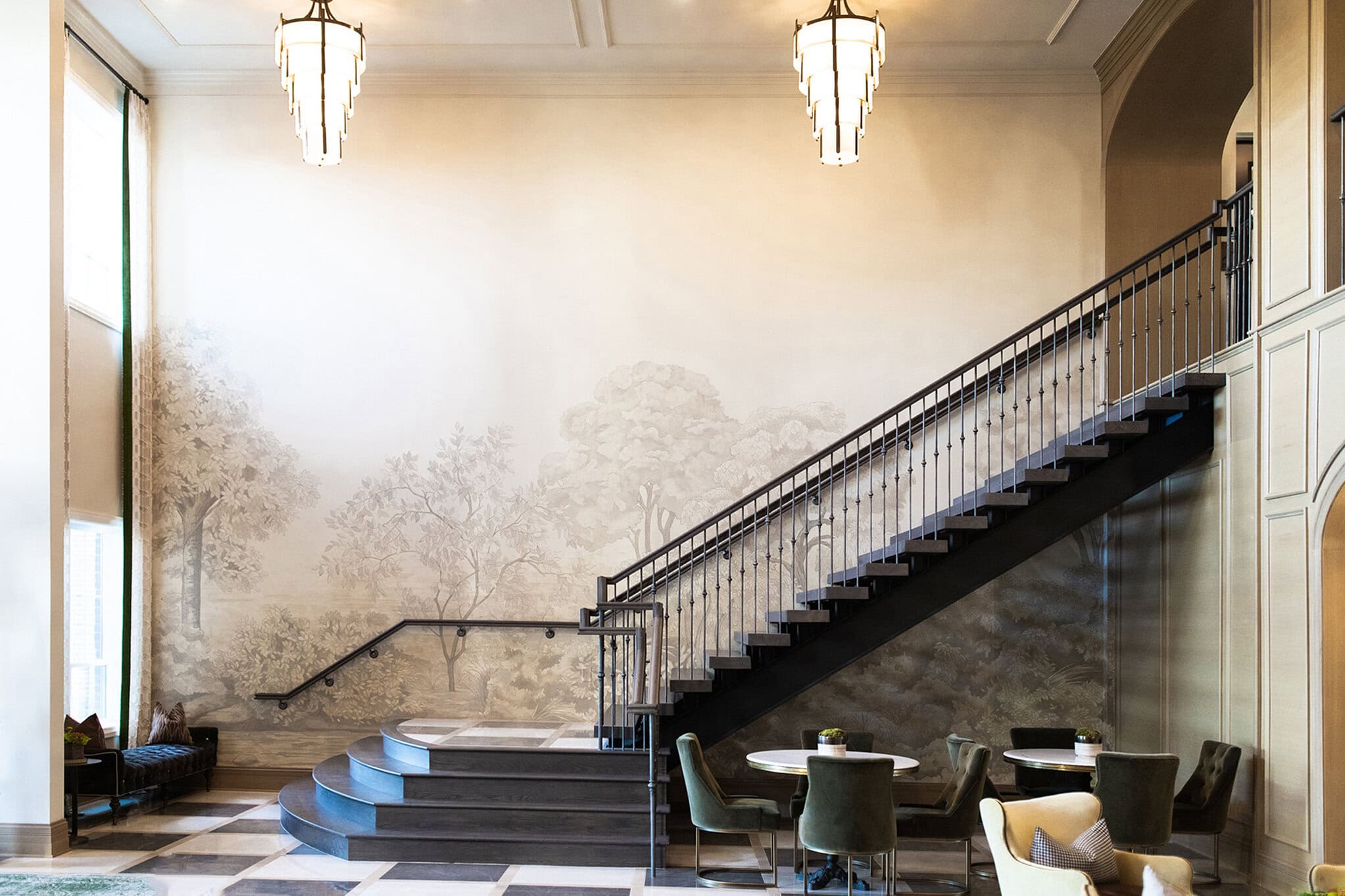 A modern staircase with dark railing rises beside a mural of trees in a spacious room with checkerboard flooring and seating areas with tables and chairs.