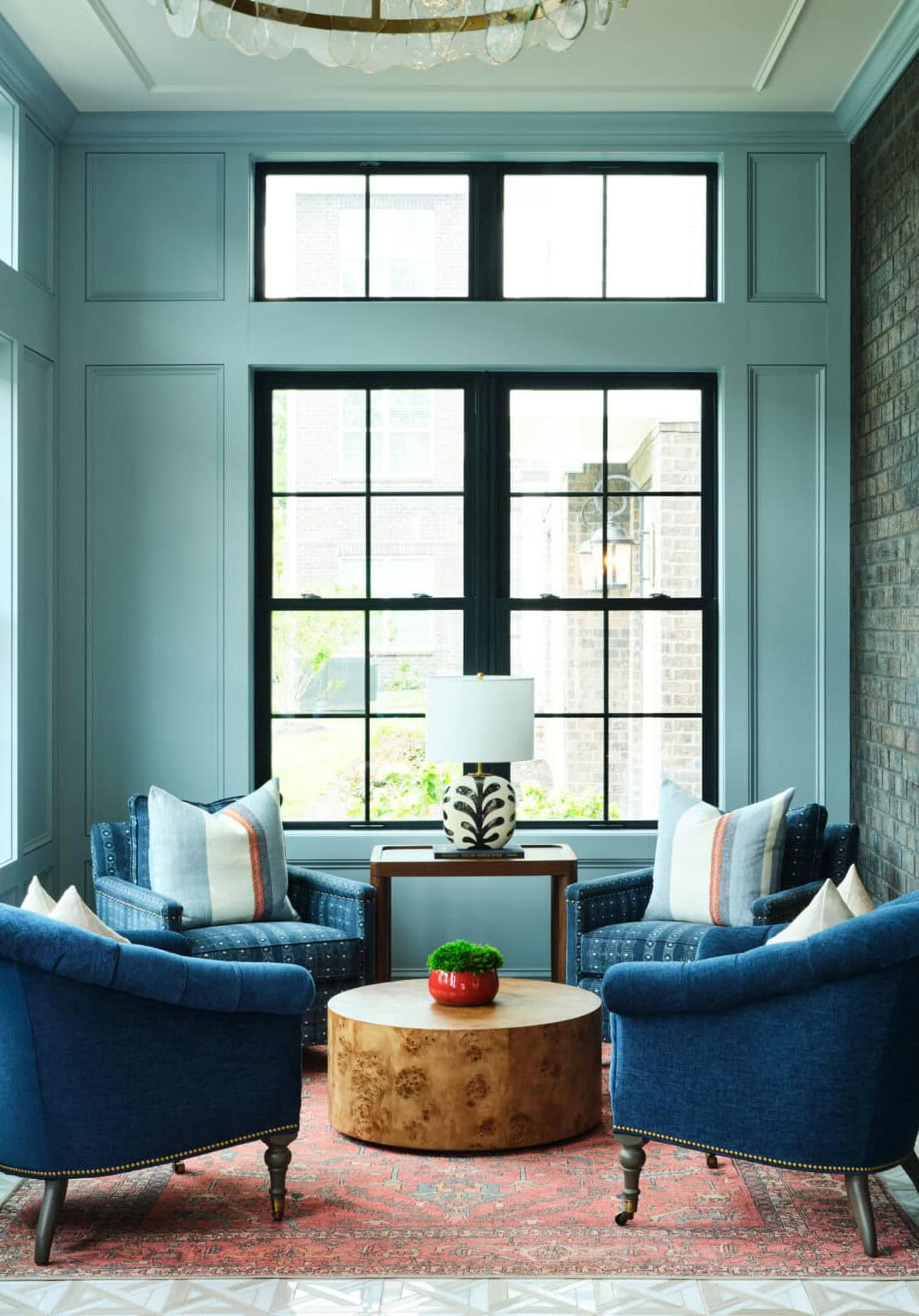 A sitting area with two blue armchairs, a round wooden coffee table, a lamp on a side table, and large windows letting in natural light.