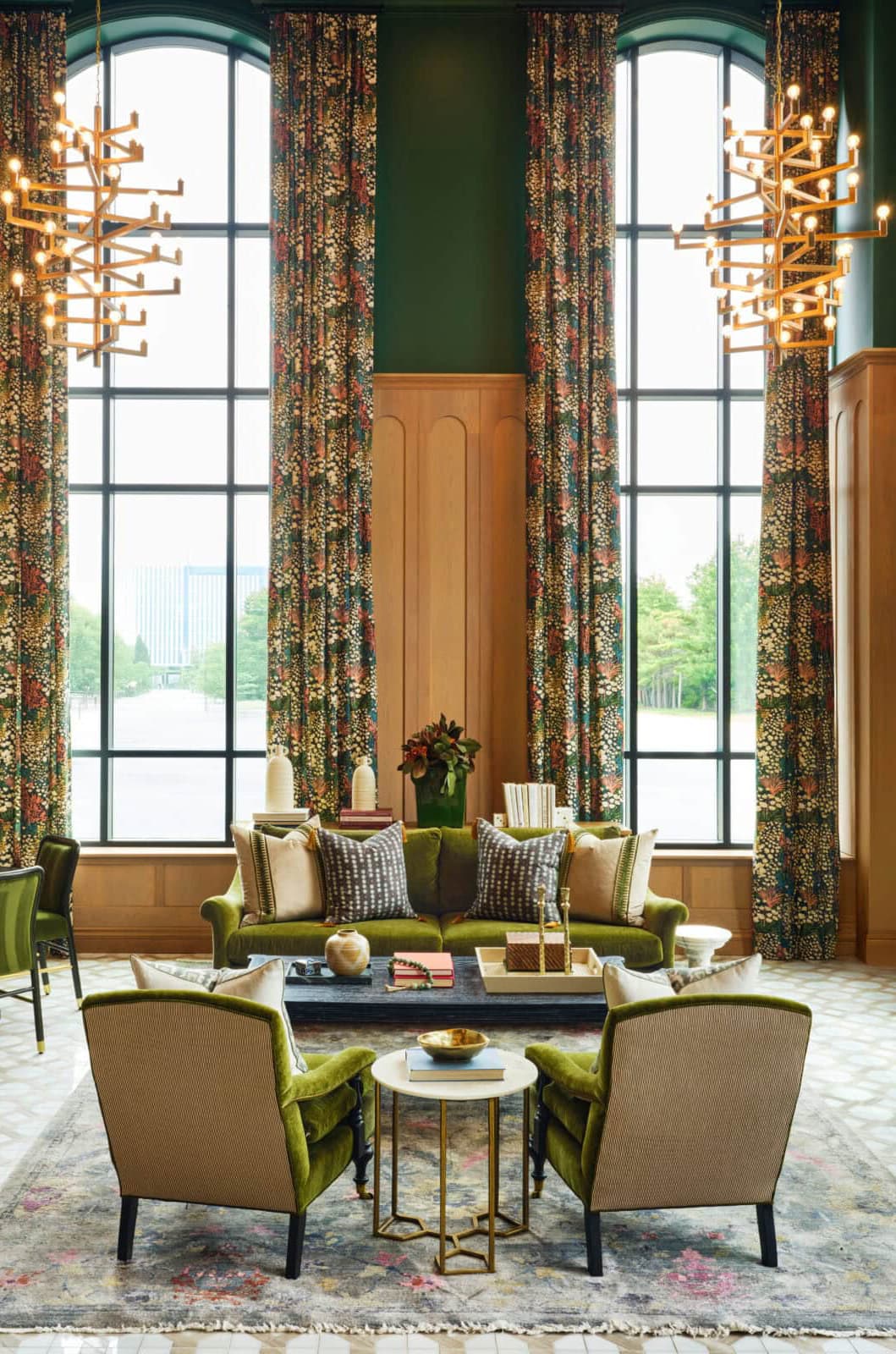 A high-ceiling multi-family amenity lobby featuring tall arched windows, deep green walls, velvet olive-green furniture, and geometric gold chandeliers.