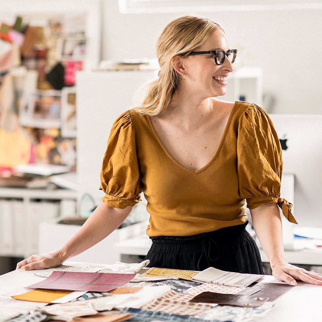 A woman wearing glasses and a mustard yellow top stands at a table covered with various fabric swatches and papers in a bright, creative workspace.