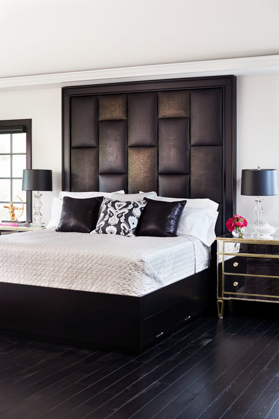 Modern bedroom with a large padded headboard, dark wood bed frame, white bedding, black and white pillows, two mirrored nightstands, lamps, and decorative items.