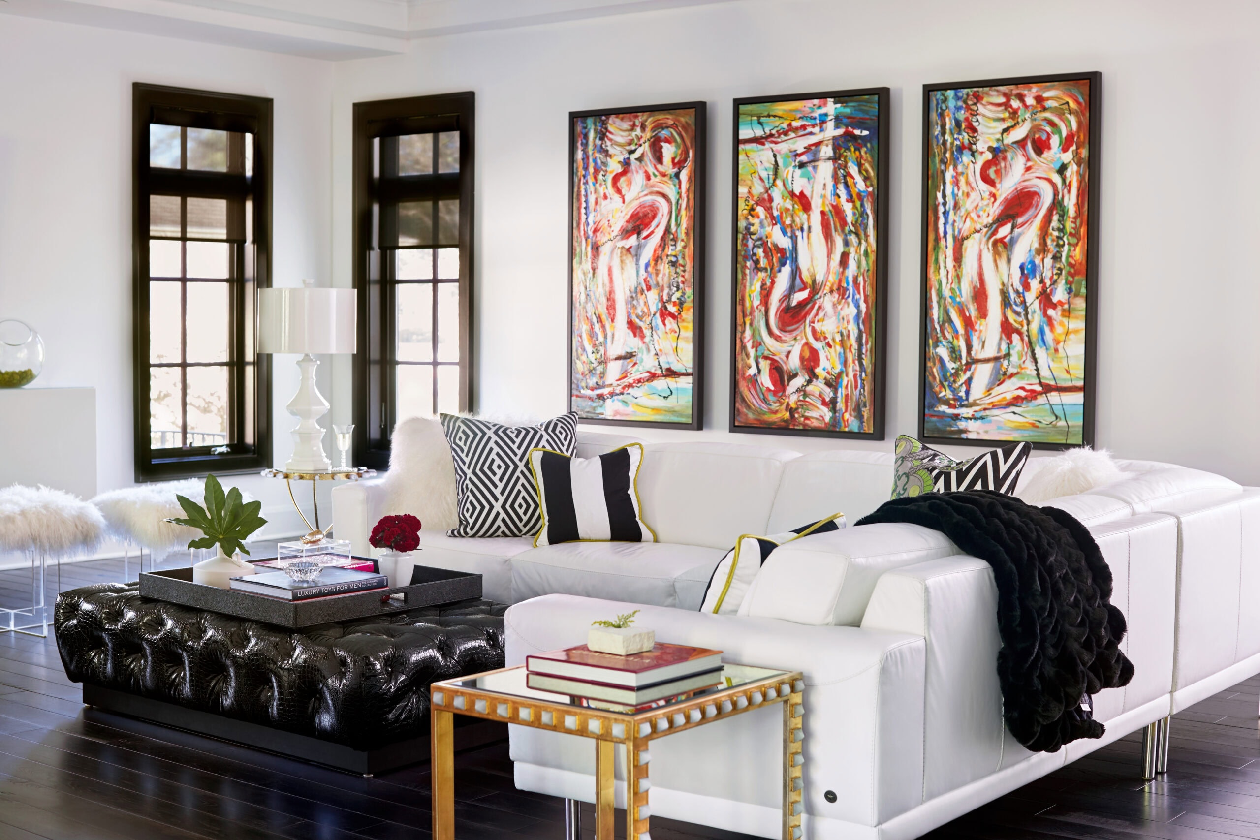 Modern living room with a white sectional sofa, black tufted ottoman, colorful abstract wall art, and decorative cushions and accessories. Large windows let in natural light.