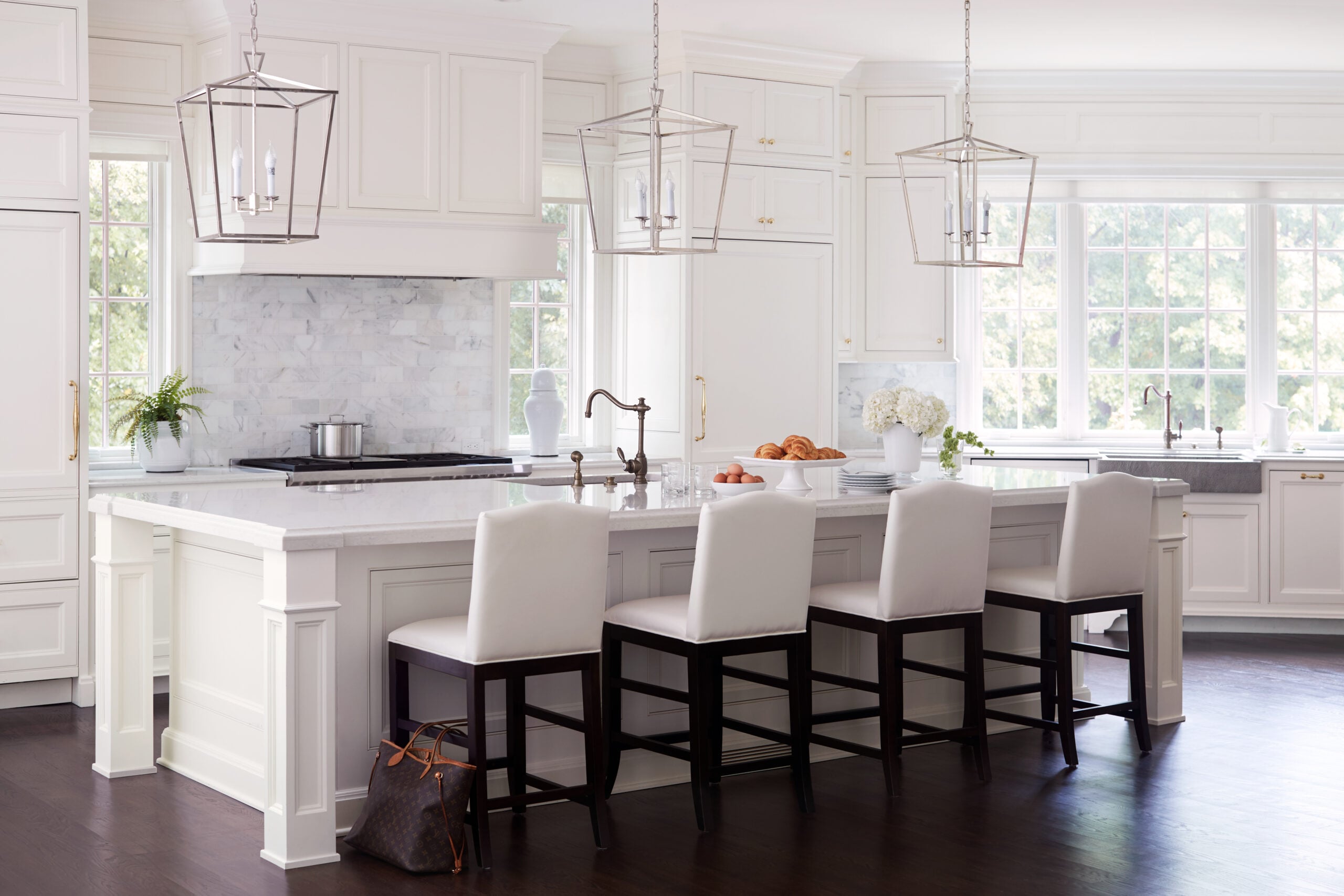 Modern white kitchen with a large island, three white cushioned chairs, pendant lights, marble countertops, and large windows letting in natural light.