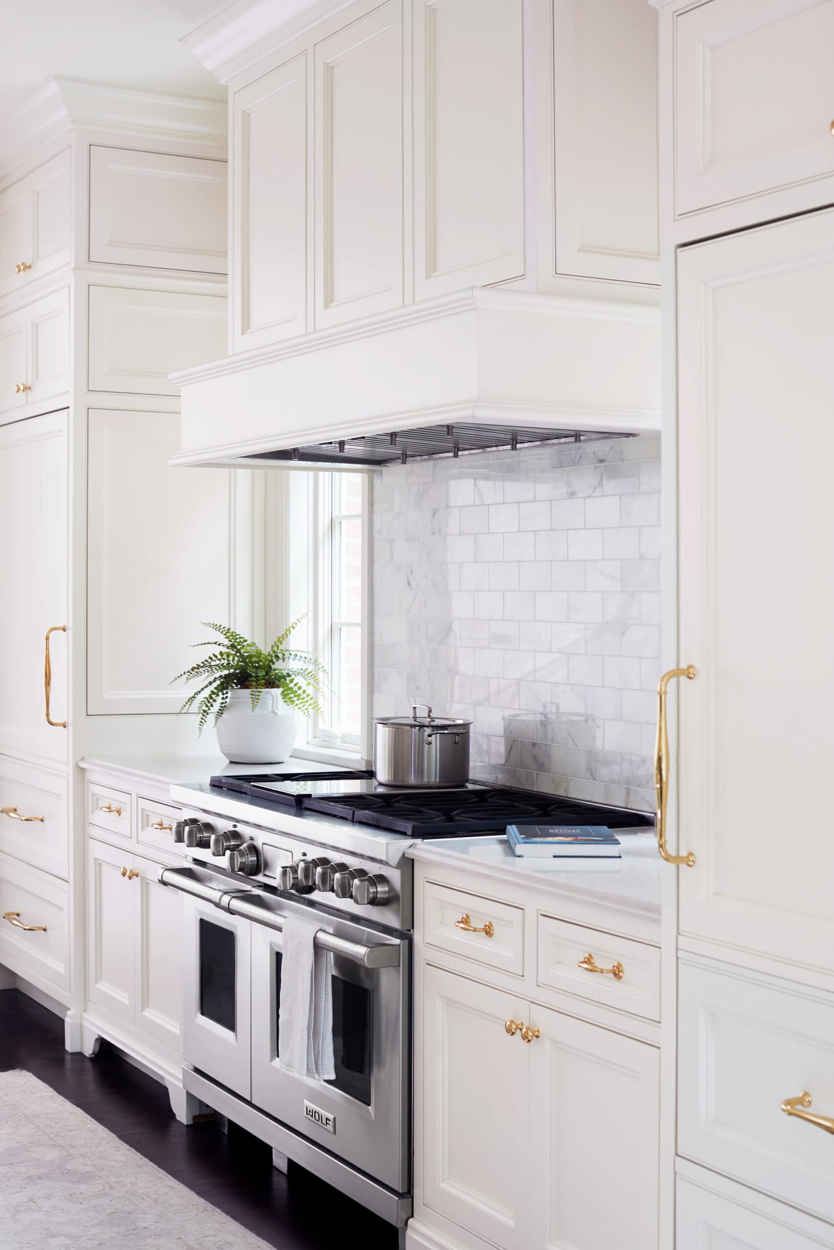 A modern kitchen with white cabinets, a stainless steel stove, a pot on the burner, a potted plant, and gold hardware. Marble backsplash and dark wood flooring are visible.