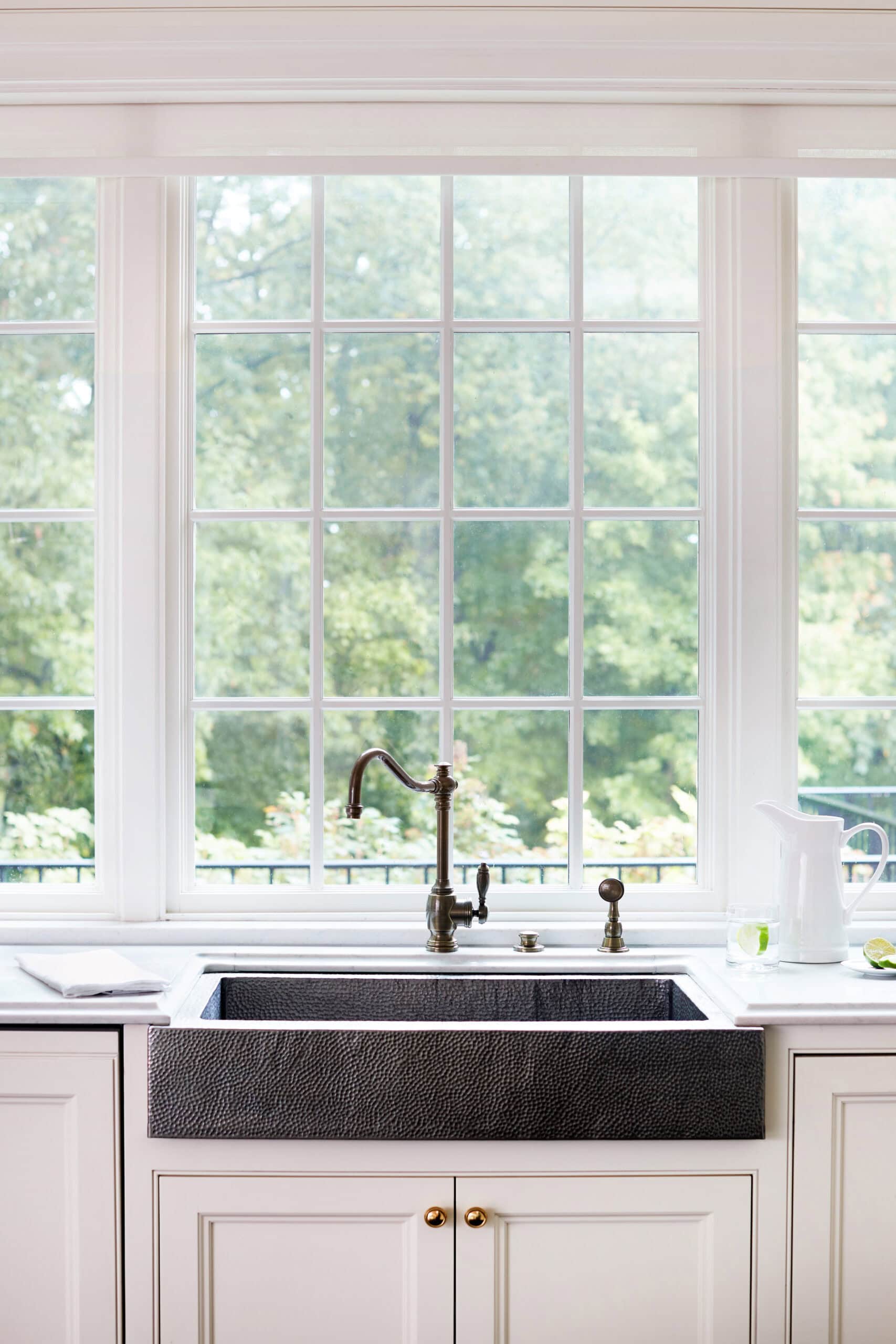 A farmhouse-style kitchen sink with a bronze faucet is set in white cabinetry beneath a large window overlooking greenery. A white pitcher and a glass sit on the counter.