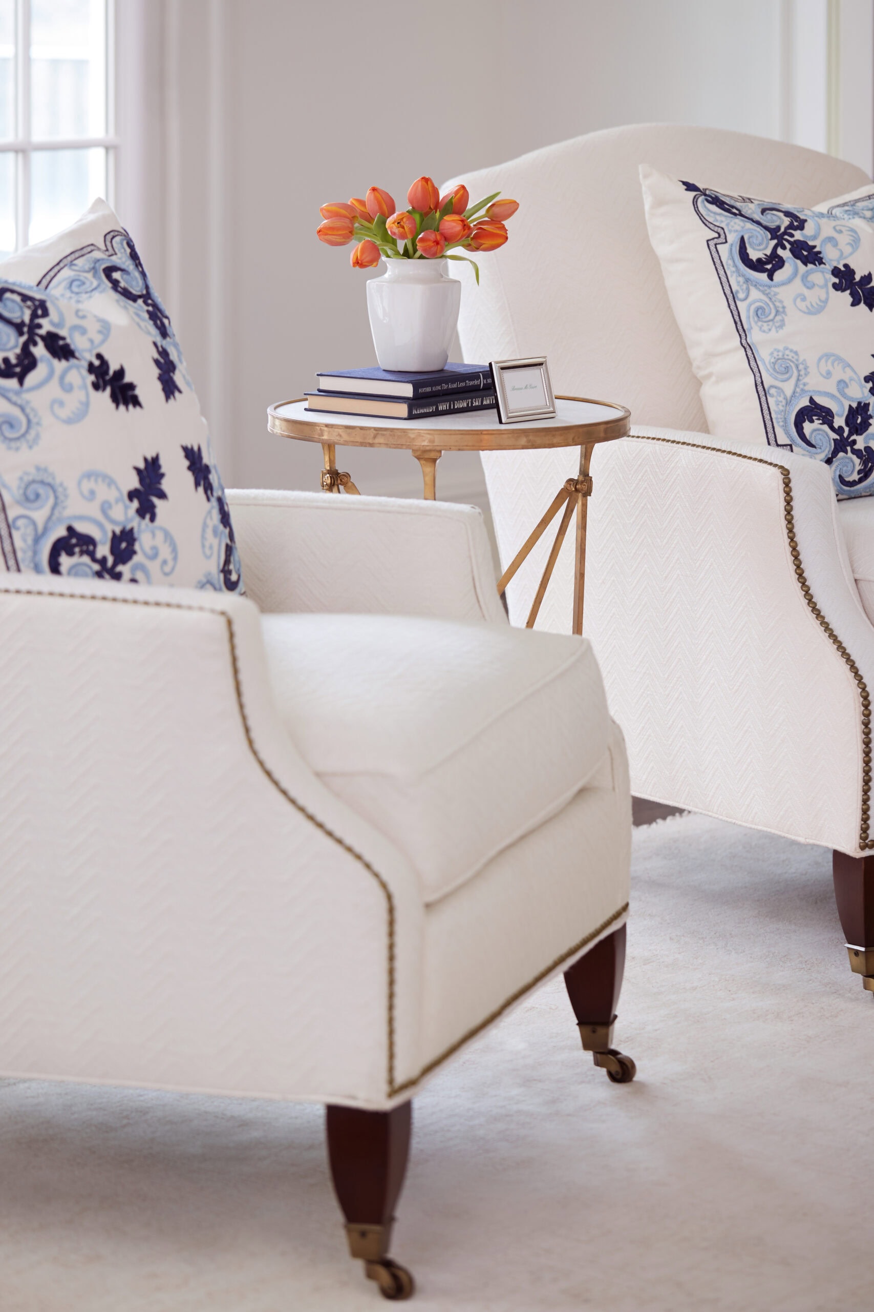 Two white armchairs with patterned blue pillows flank a small gold side table holding books, a framed photo, and a vase of orange tulips in a bright, softly-lit room.
