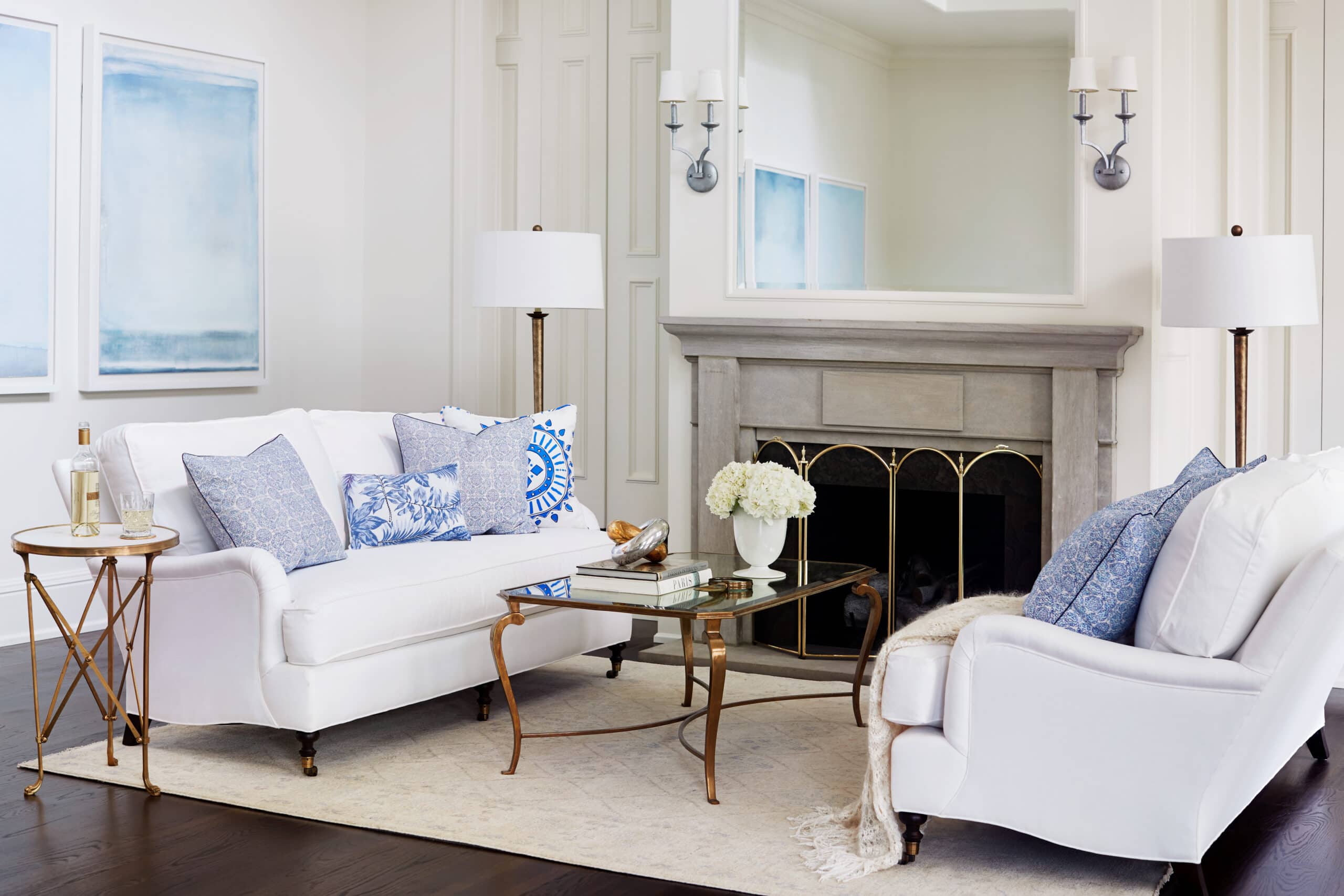 Bright living room with two white sofas, blue patterned pillows, glass coffee table, gray fireplace, wall mirrors, and white lamps on either side. Two abstract paintings hang on the wall.