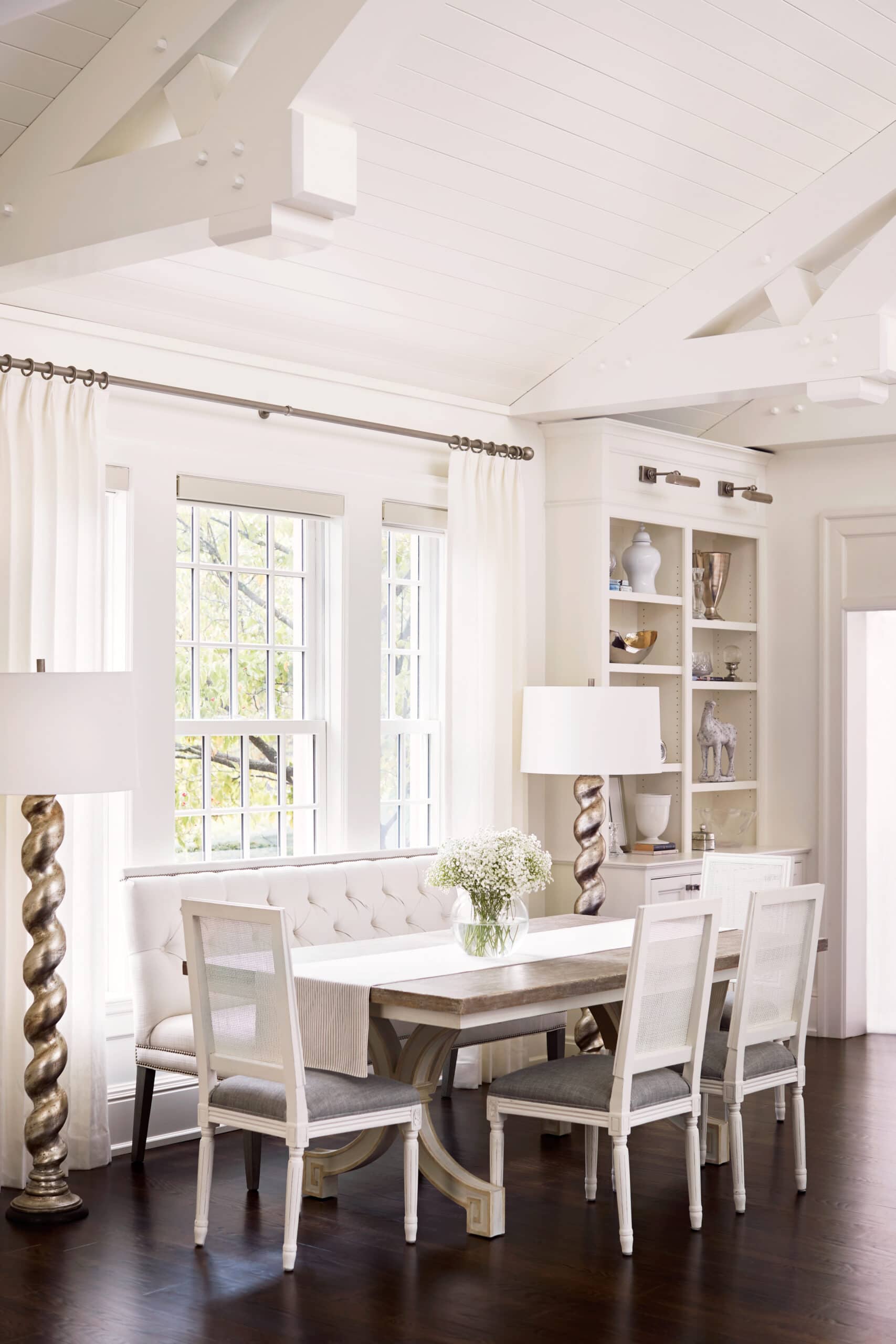 A bright dining area with white furniture, a rectangular table, four chairs, a bench, two floor lamps, shelves with decor, and large windows with white curtains.