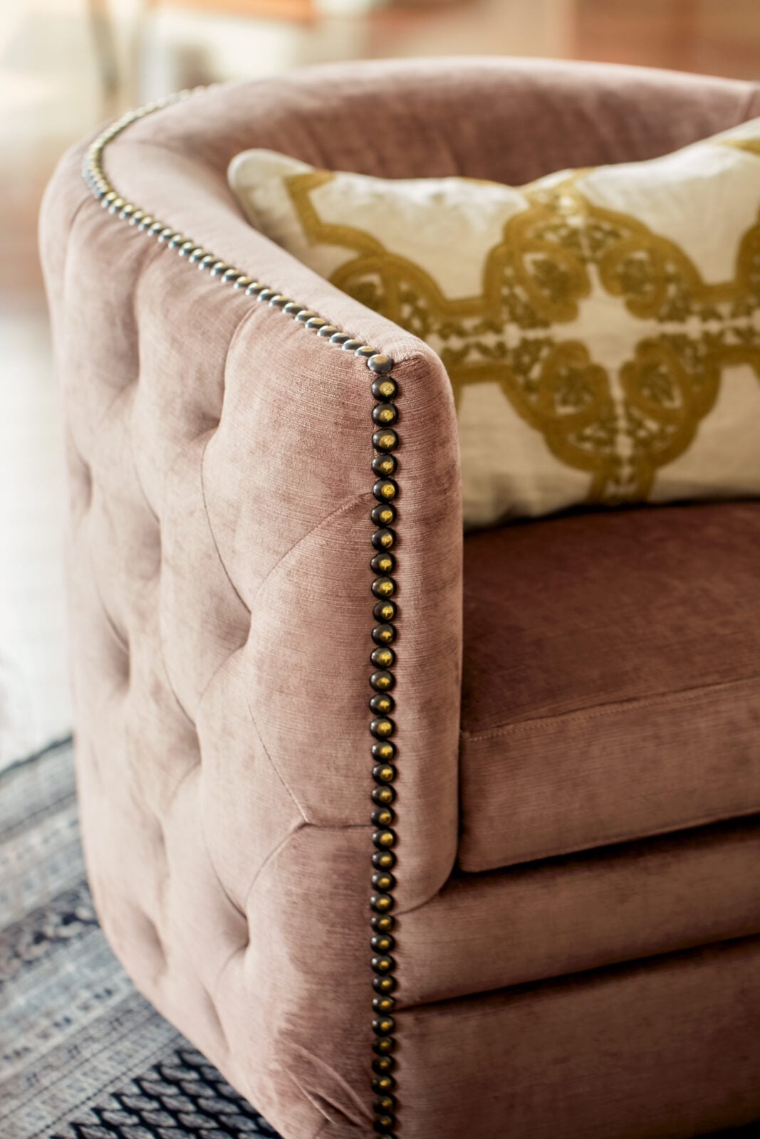 Close-up of a mauve tufted armchair with brass stud detailing and a cream decorative pillow with an olive green pattern.