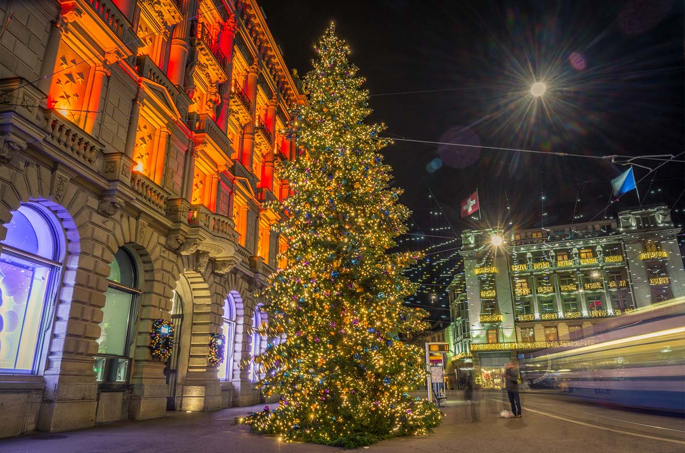 Image of Christmas tree lit up at nighttime in Switzerland. Buildings alongside the tree are lit up with a warm glow and look welcoming and have an ambient atmosphere