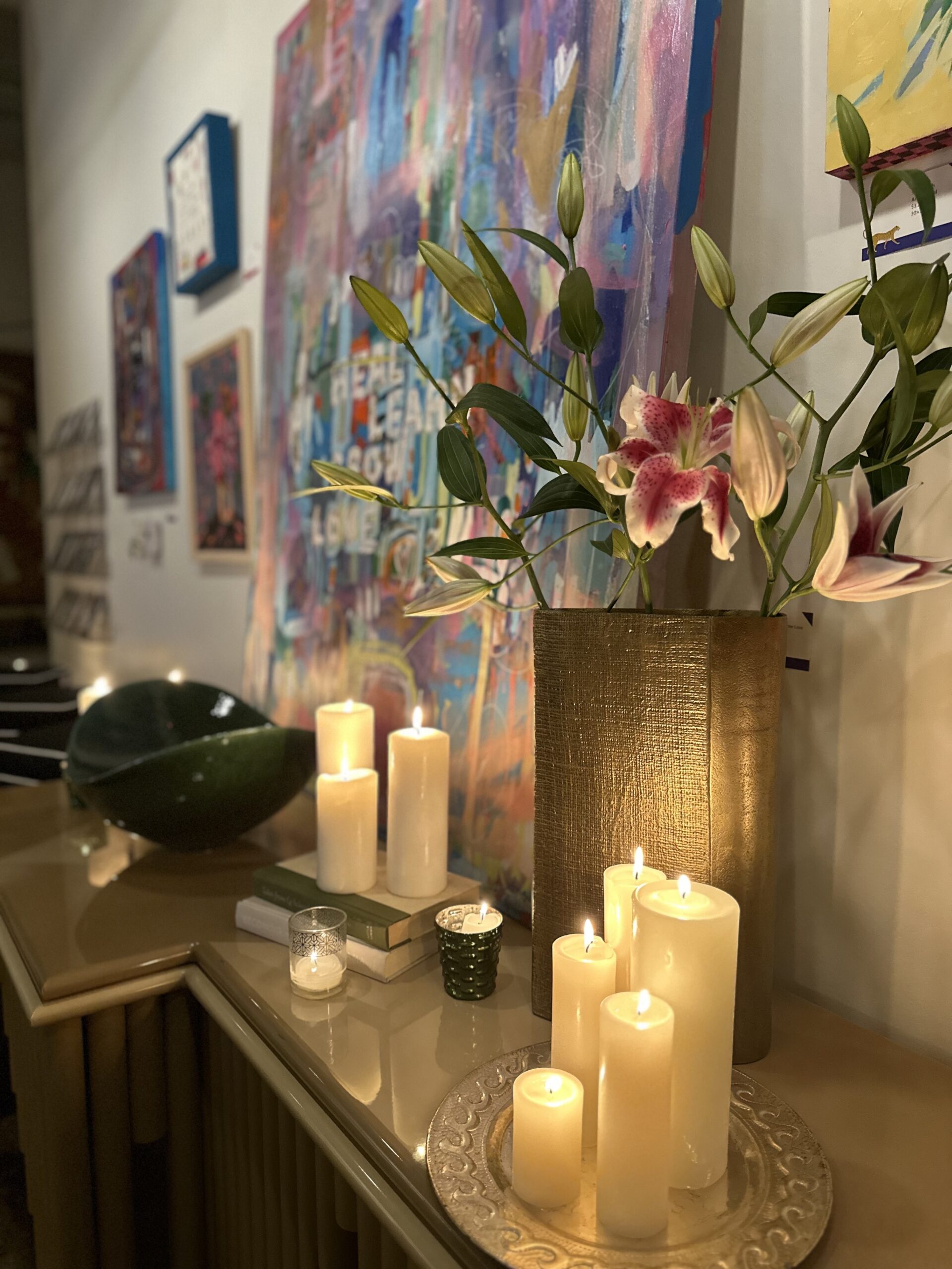 Candles dimly lit all over a table, lighting up a white and pink orchid as well as pastel and colorful artwork in background