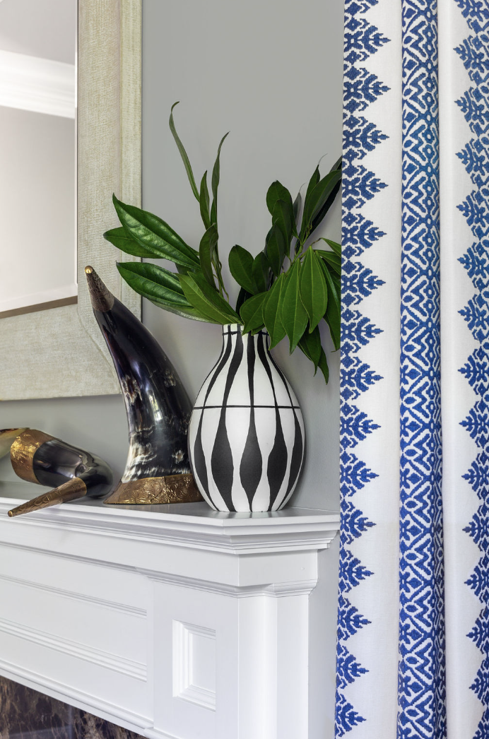 Black and white patterned vase on mantle with greenery in the vase. Blue and white accents on wall near the vase. Gray background walls.