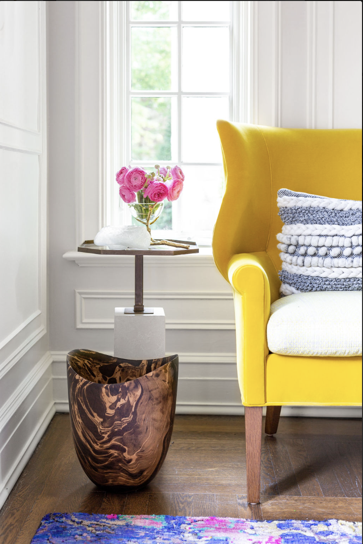 Yellow sitting chair in living room with throw pillow sitting on it. With a decorative side table with pink flowers in a vase on it, with a wooden trash bin for decoration below.