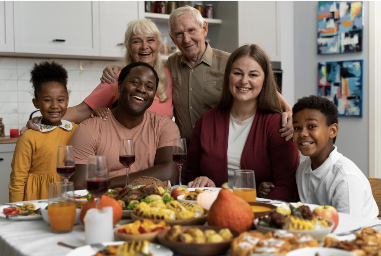 Family of different races all sitting around a kitchen table together sharing a meal. All different generations are photographed, everyone is smiling and looking at the camera