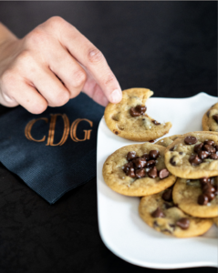 Fresh hot plate of chocolate chip cookies on a white plate with a black embellished napkin. A hand is grabbing pointer fingers to pick up the cookies.