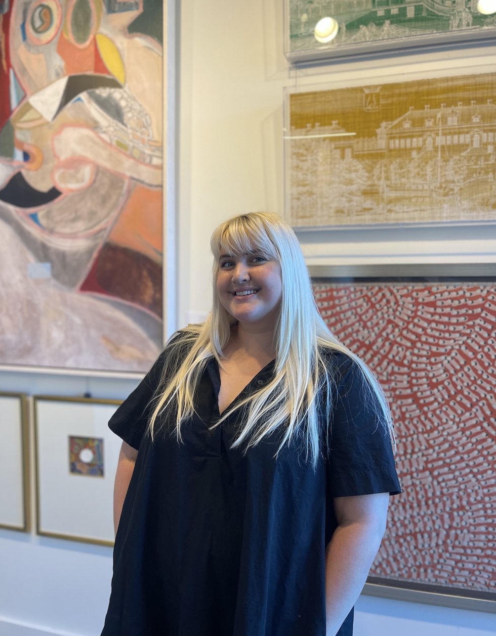 New employee headshot. Woman with long blonde and bright hair and bangs stands in front of artwork that is colorful and bright