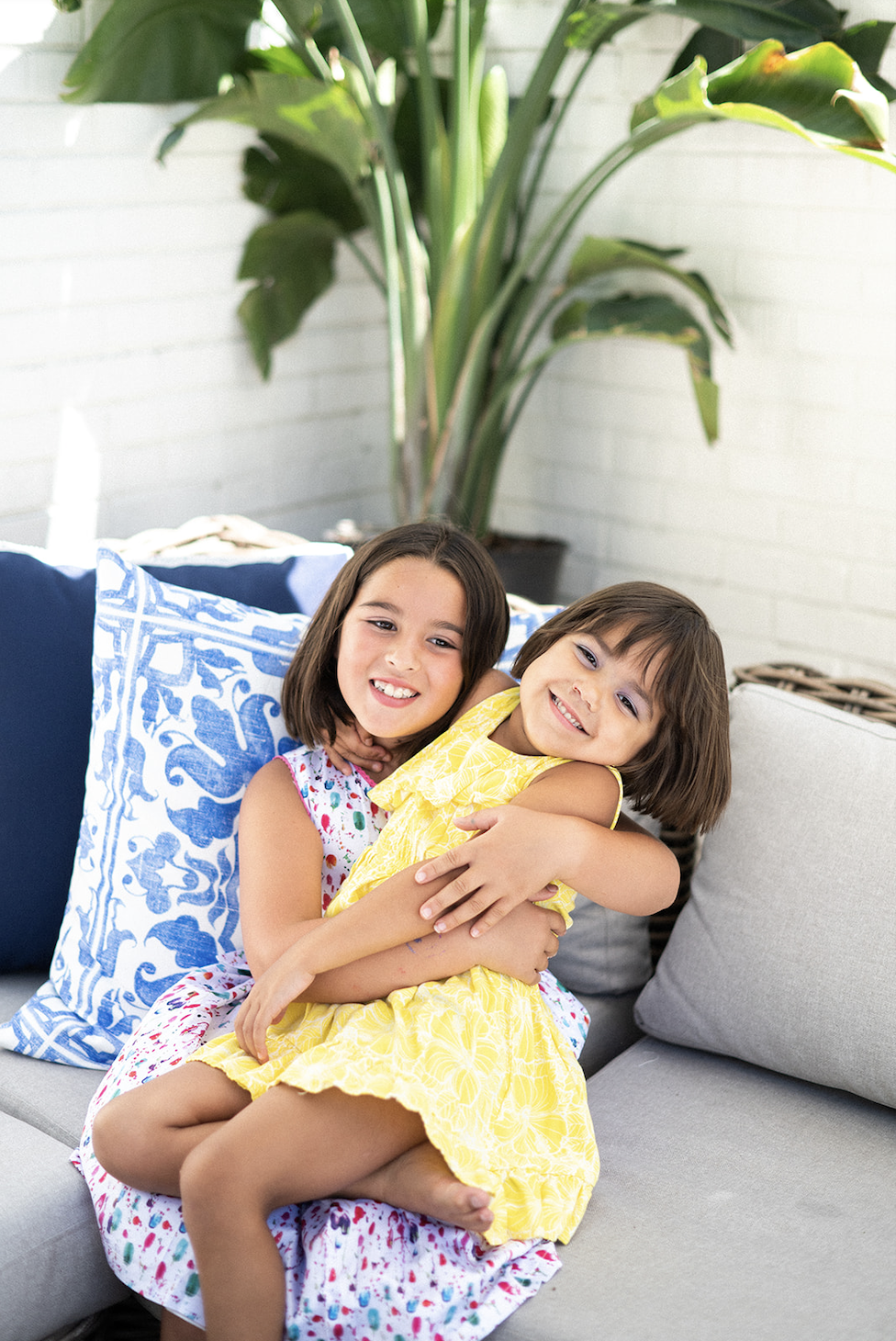 two young girls that are sisters sitting on an outdoor sofa with live plants in the background, hugging one another