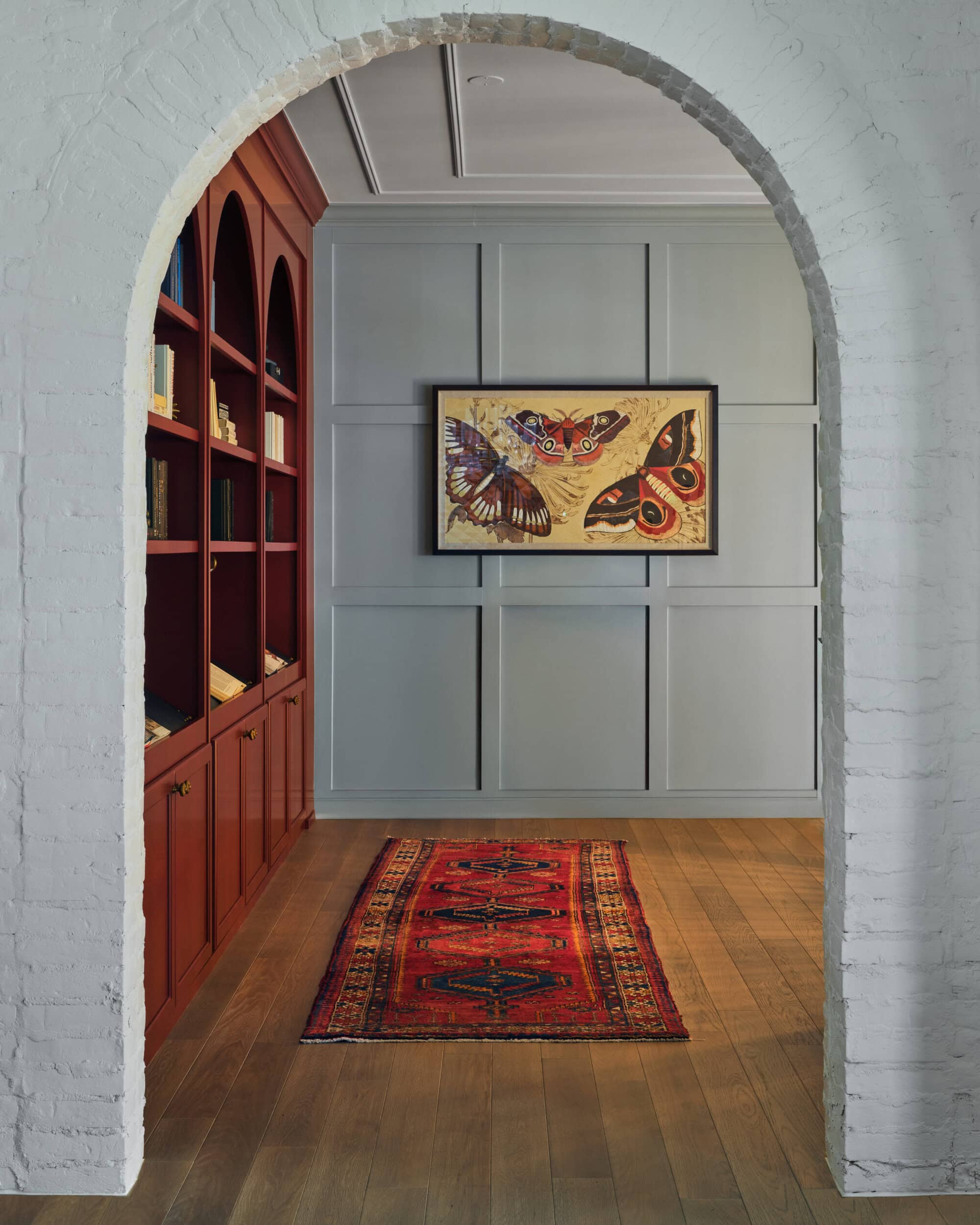 View through a white brick archway of a hallway with a red rug, red built-in shelves on the left, paneled gray wall, and a framed butterfly artwork.
