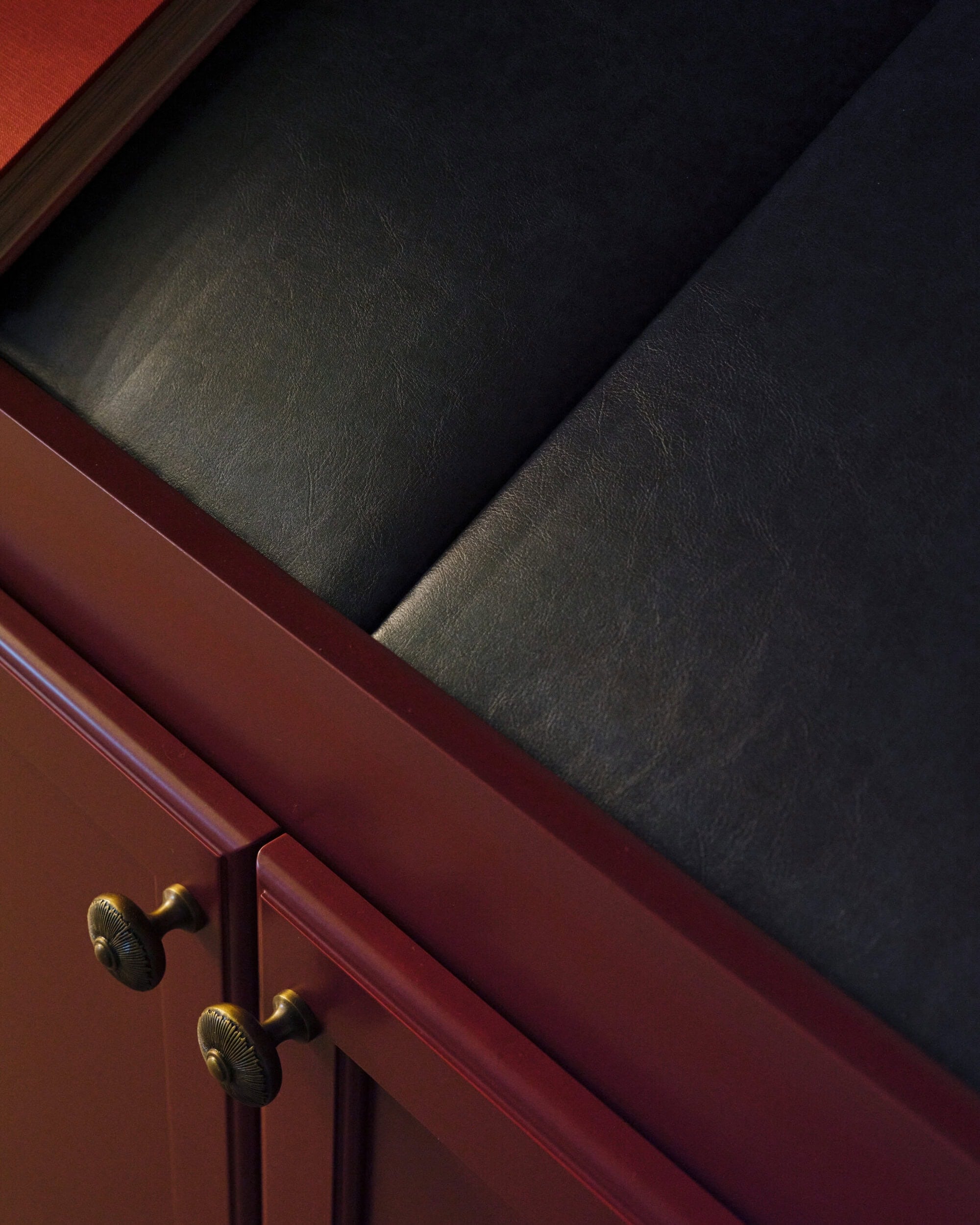 Close-up of a red cabinet with brass handles and a black cushioned seat on top, showing the textures of wood and leather-like material.