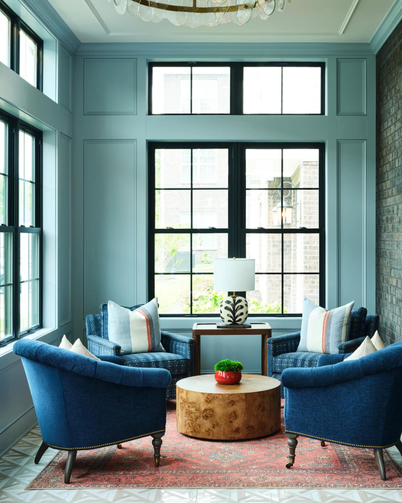 A sunlit sitting area with four blue armchairs, a round wooden coffee table, a small lamp, and a red rug near large windows and a brick accent wall.