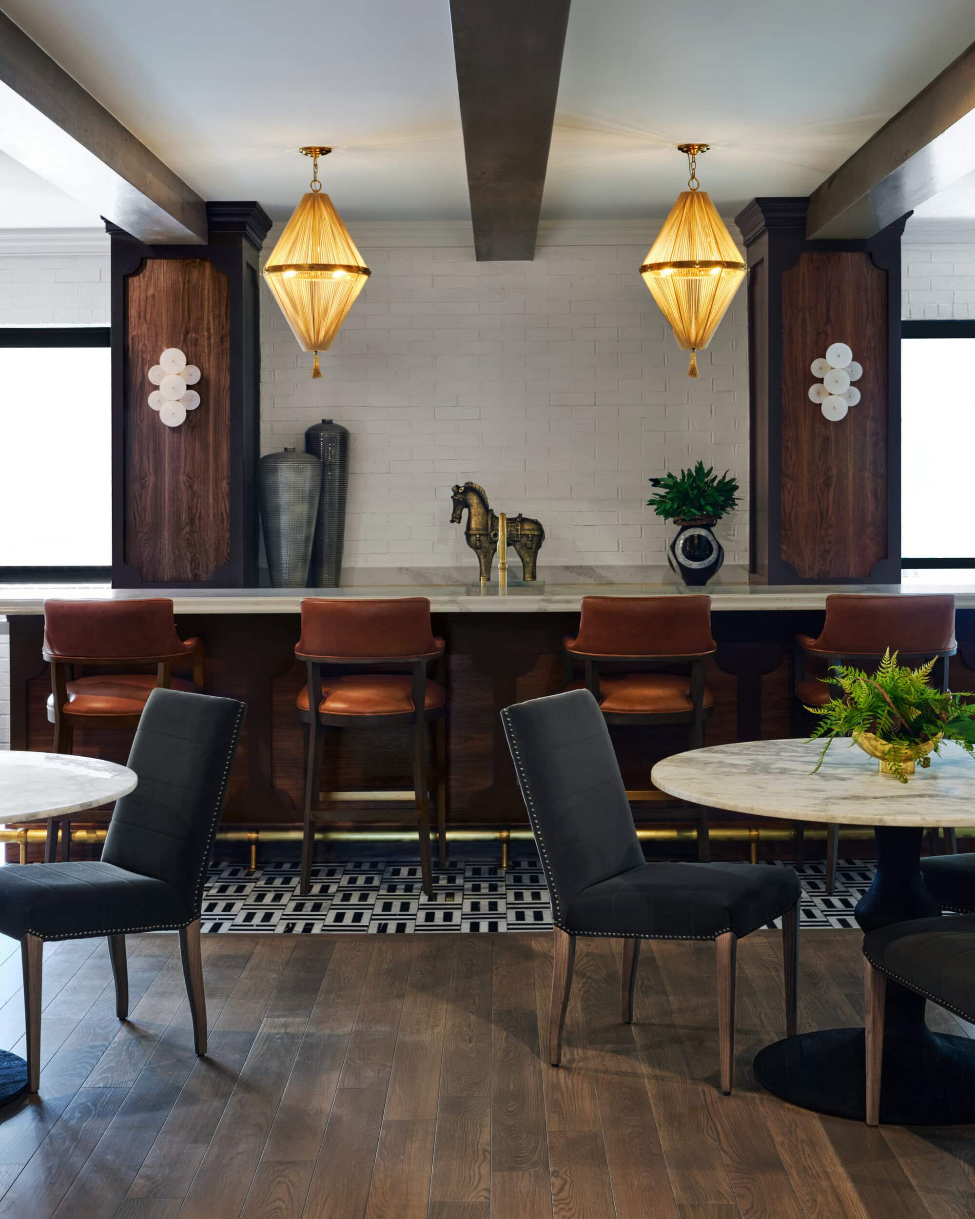 A modern bar area with brown leather barstools, a marble countertop, gold horse statues, and geometric pendant lights above. Black dining chairs and round tables are in the foreground.