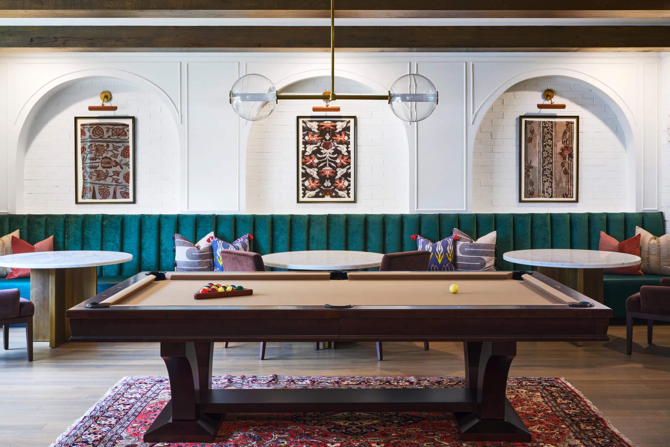 A pool table sits on a patterned rug in front of green upholstered booth seating with round tables, framed artwork, and pendant lighting in an elegant room.