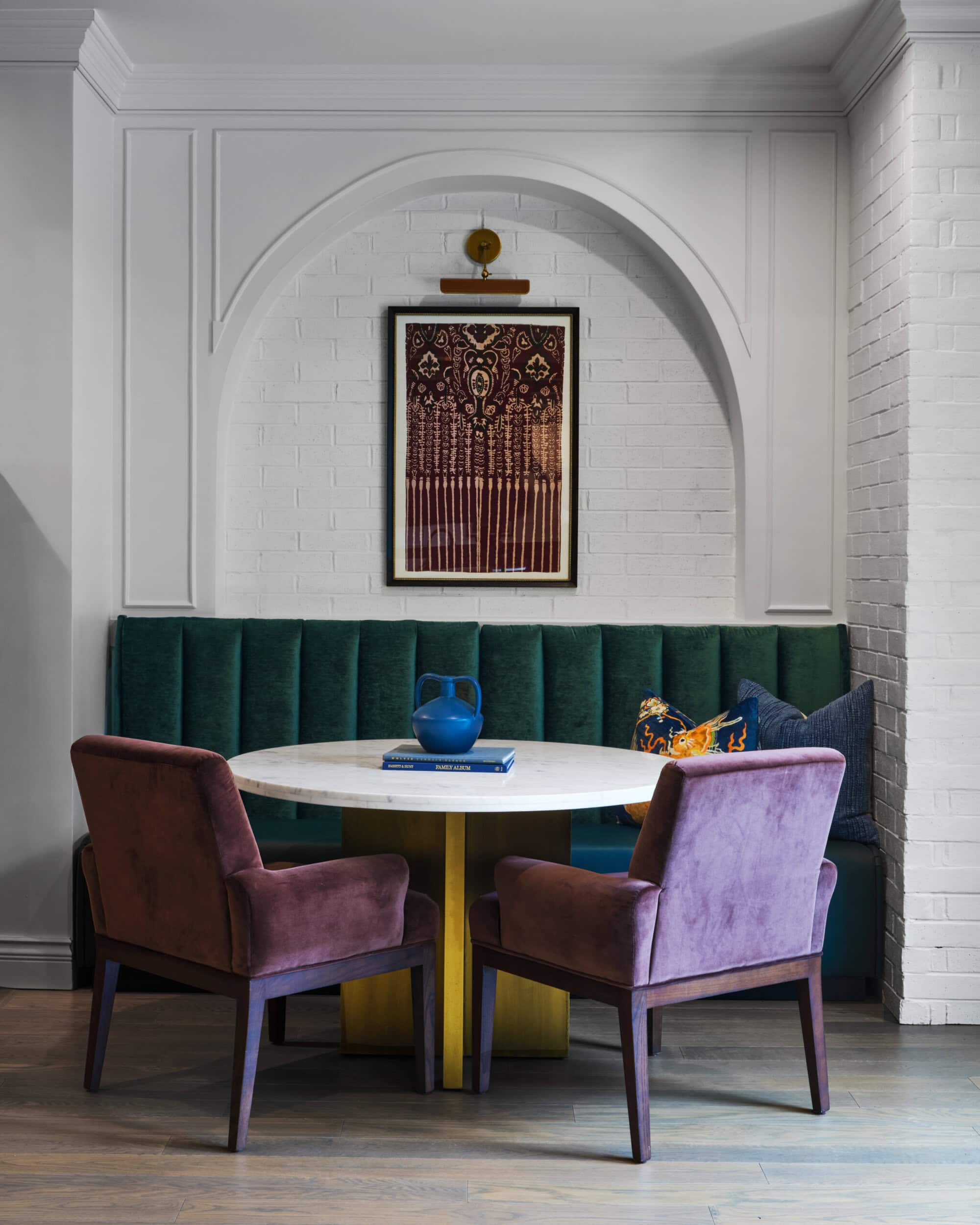 A round marble table with two purple chairs and a green velvet banquette sits below a framed artwork in a white-paneled alcove.