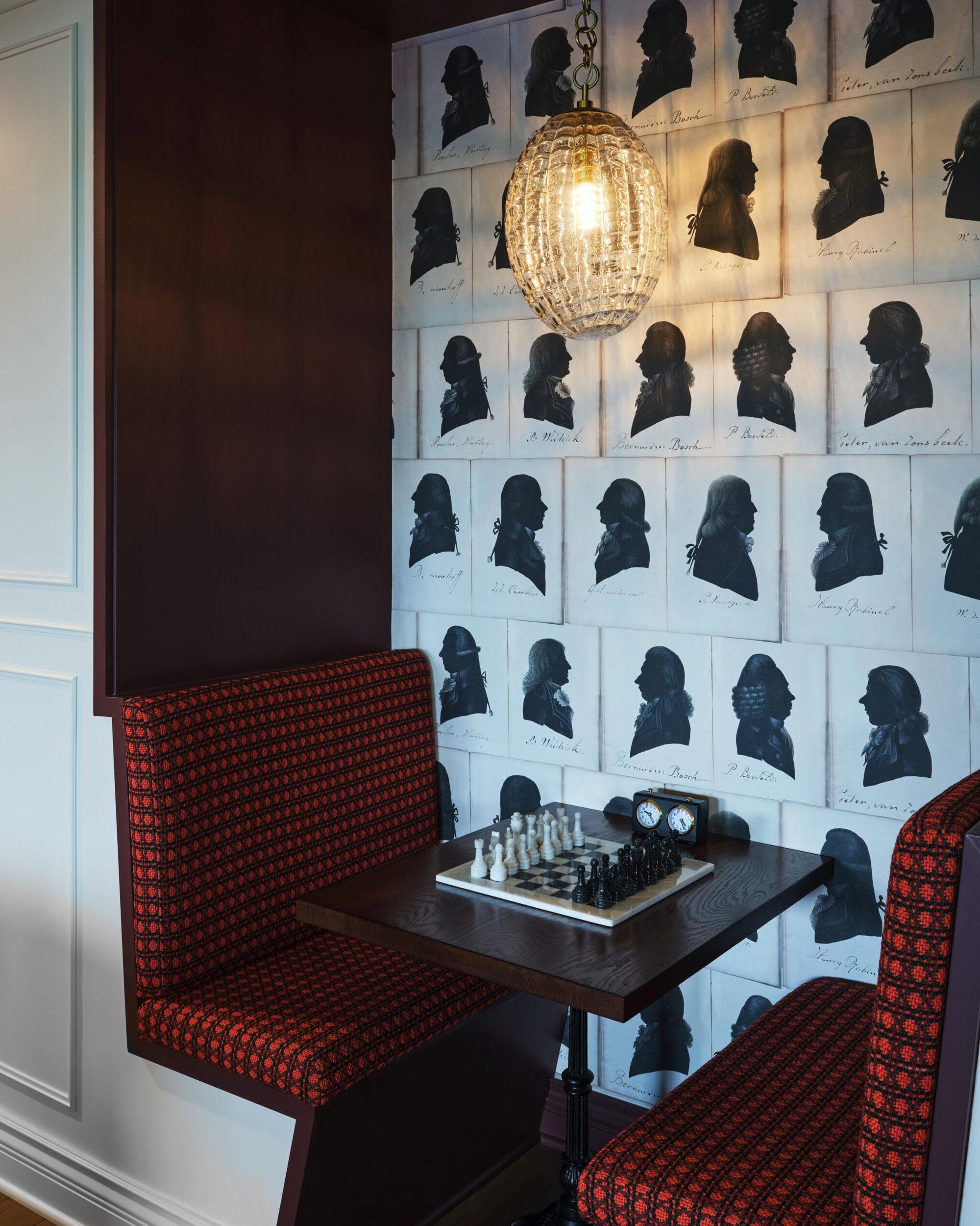 A small seating nook with a chessboard on a table, red upholstered benches, a hanging glass light fixture, and wallpaper featuring black and white silhouette portraits.