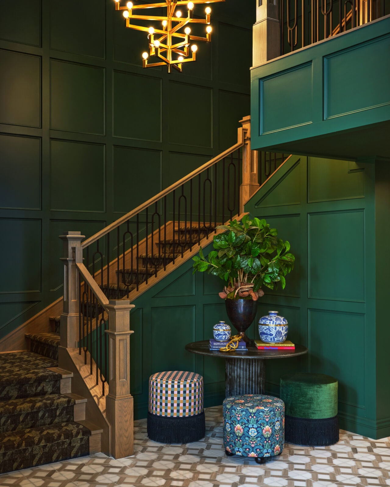 A staircase with wooden handrails, dark green paneled walls, a geometric light fixture, a round table with vases, a potted plant, and colorful upholstered stools.