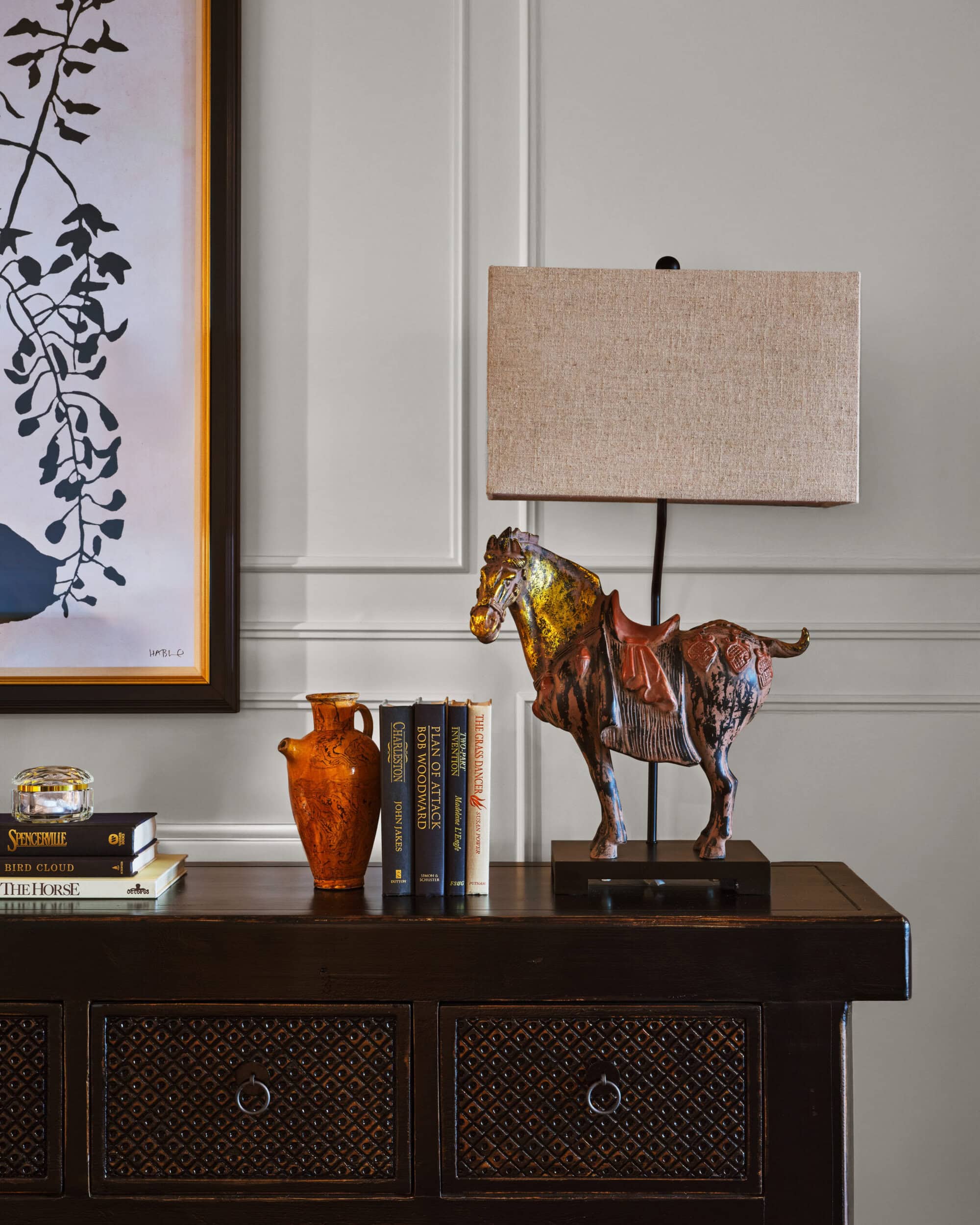 A dark wood console table with decorative books, an amber vase, a horse-shaped lamp with a beige shade, and wall art partially visible in the background.