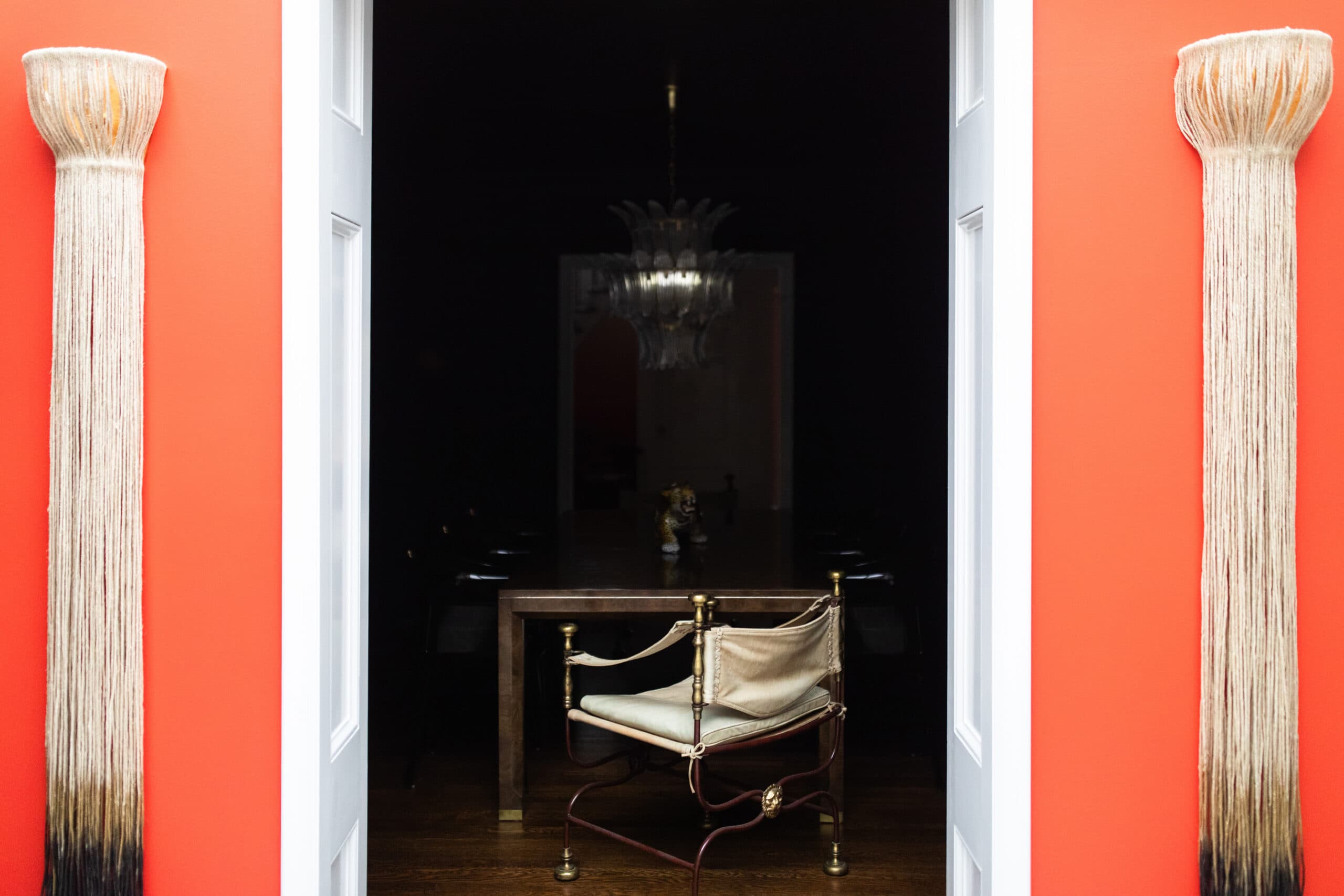 A brass and leather chair sits at a wooden table in a dark room, framed by two white doors and bright red walls with tasseled wall decorations.