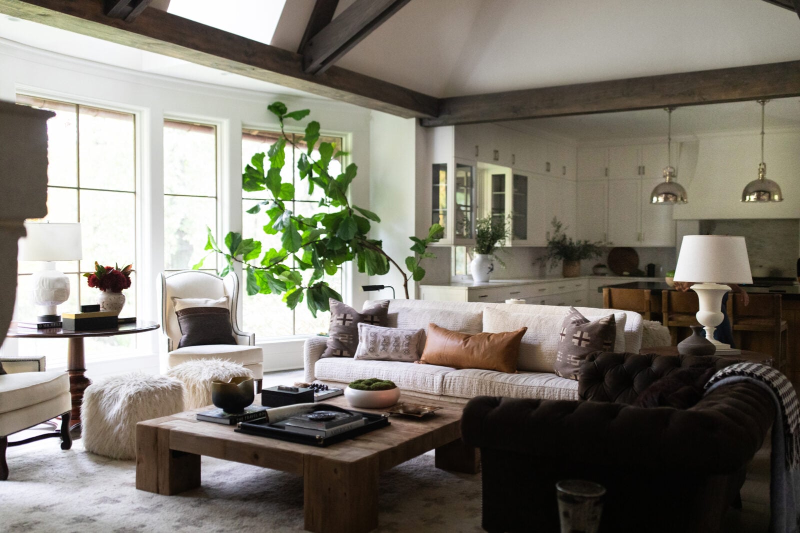 A living room with a beige sofa, a wooden coffee table, a dark armchair, large windows, indoor plants, and an open kitchen in the background.