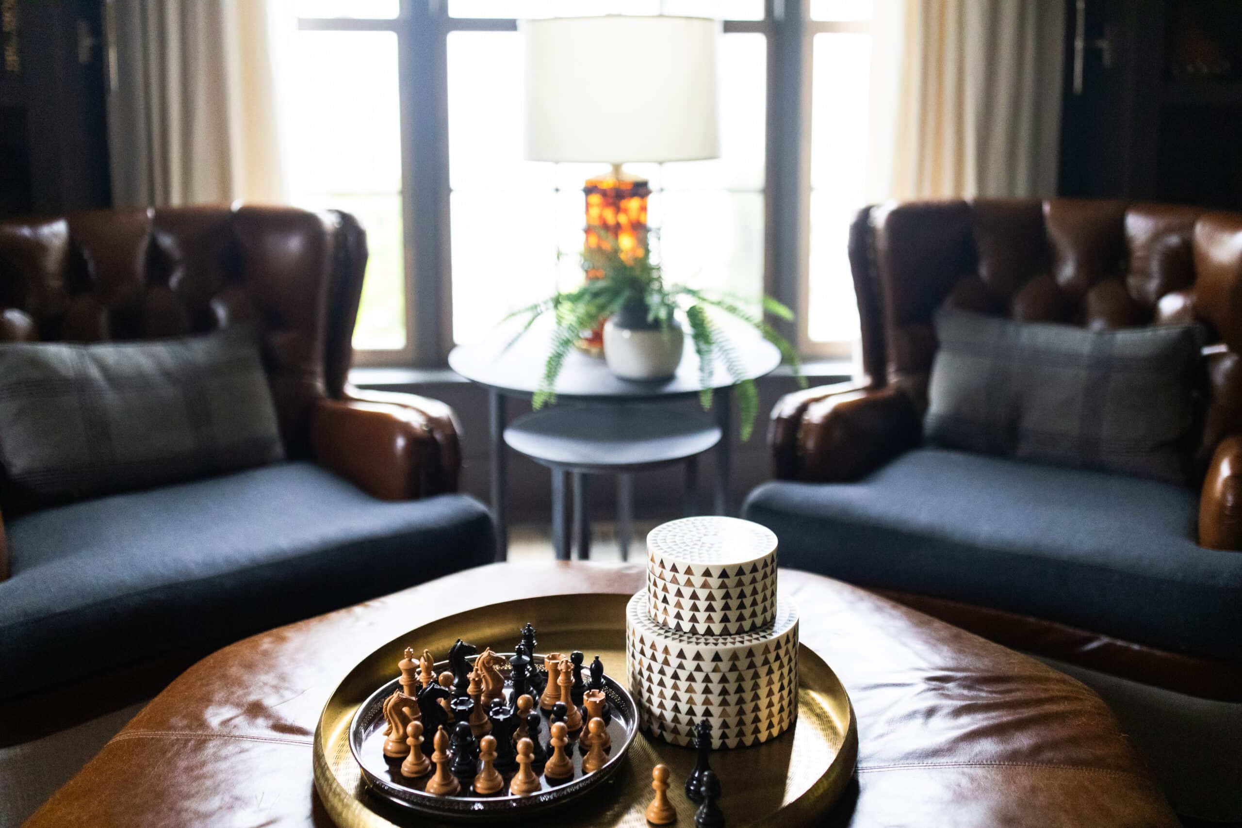 Two leather armchairs sit by a window with a round table holding a lamp and plant between them. A tray with a chess set and patterned boxes is on a leather ottoman in the foreground.