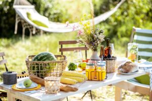 outdoor picnic scene with basket of watermelon and various fruits and vegetables, floral arrangements, jars of beverages, and a table with wooden chairs
