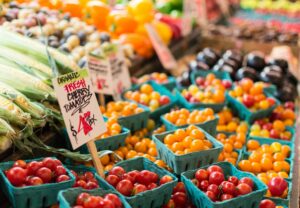 A wooden table at a farmers market showing blue baskets full of various vegetables including tomatoes, bell peppers, and corn, with small paper signs.