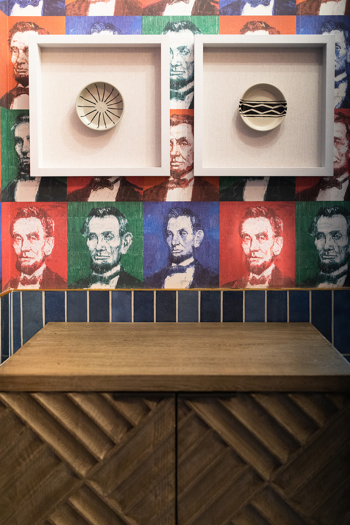 Wall with dark blue subway tile backsplash on bottom half and pop art style wallpaper of Abe Lincoln on top with two globally inspired art pieces of neutral colored circle artisan crafts with a wooden credenza cabinet beneath