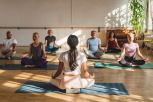 A yoga room with 7 people doing a sitting yoga pose on yoga mats and a wooden floor.
