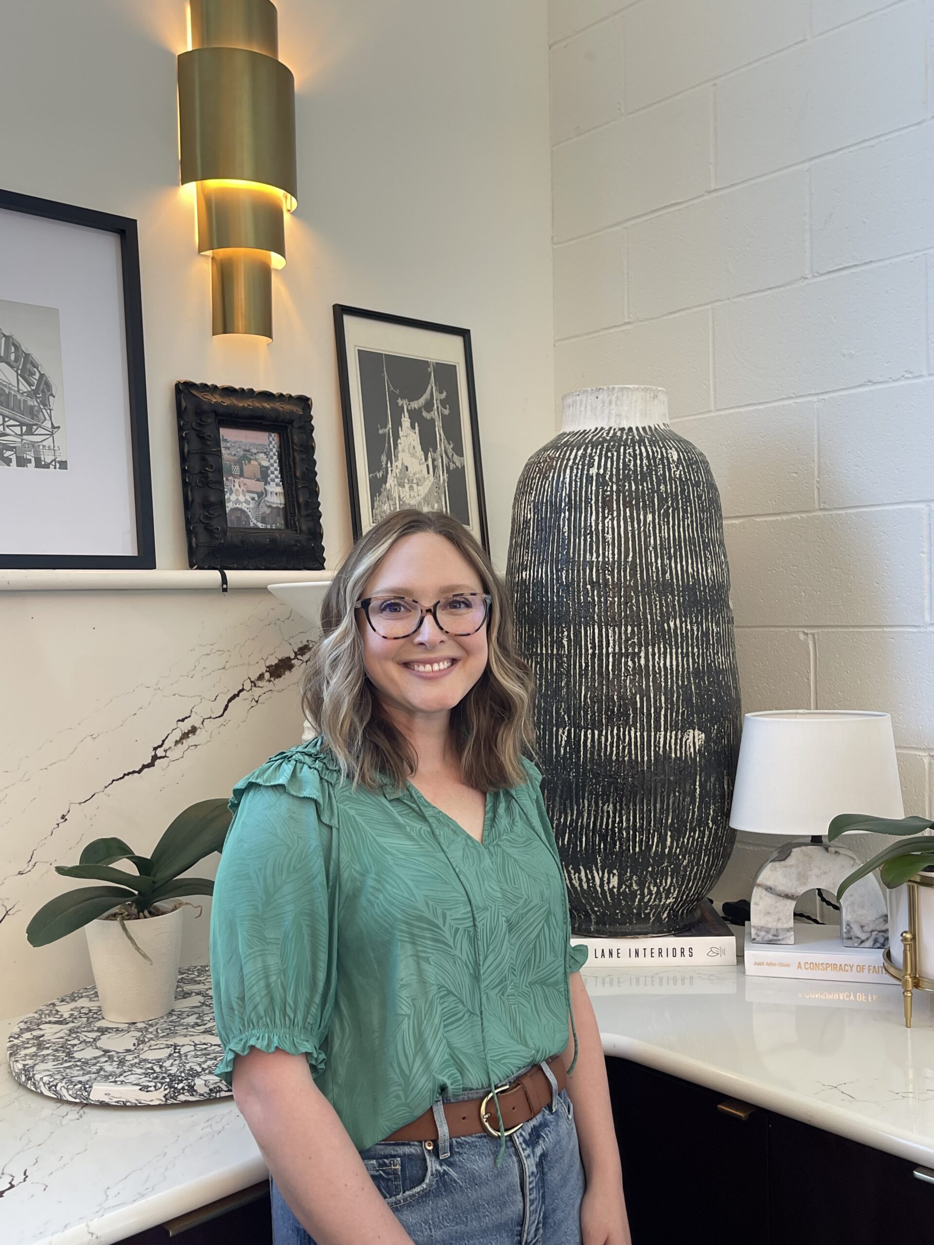 Caucasian woman with short blonde hair and black glasses wearing a teal colored blouse and blue jeans standing in front of a kitchen counter decorated with plants, a white lamp, a grey tall vase, and a black and white art gallery
