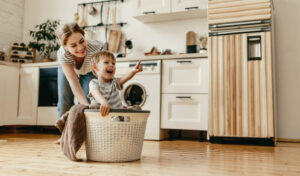 A mother and a son cleaning in a neutral colored laundry room. The mother is wearing a light grey tshirt and jeans and is bending down to hold onto a white laundry basket. The son is wearing a grey and white shirt and is sitting inside the laundry basket.