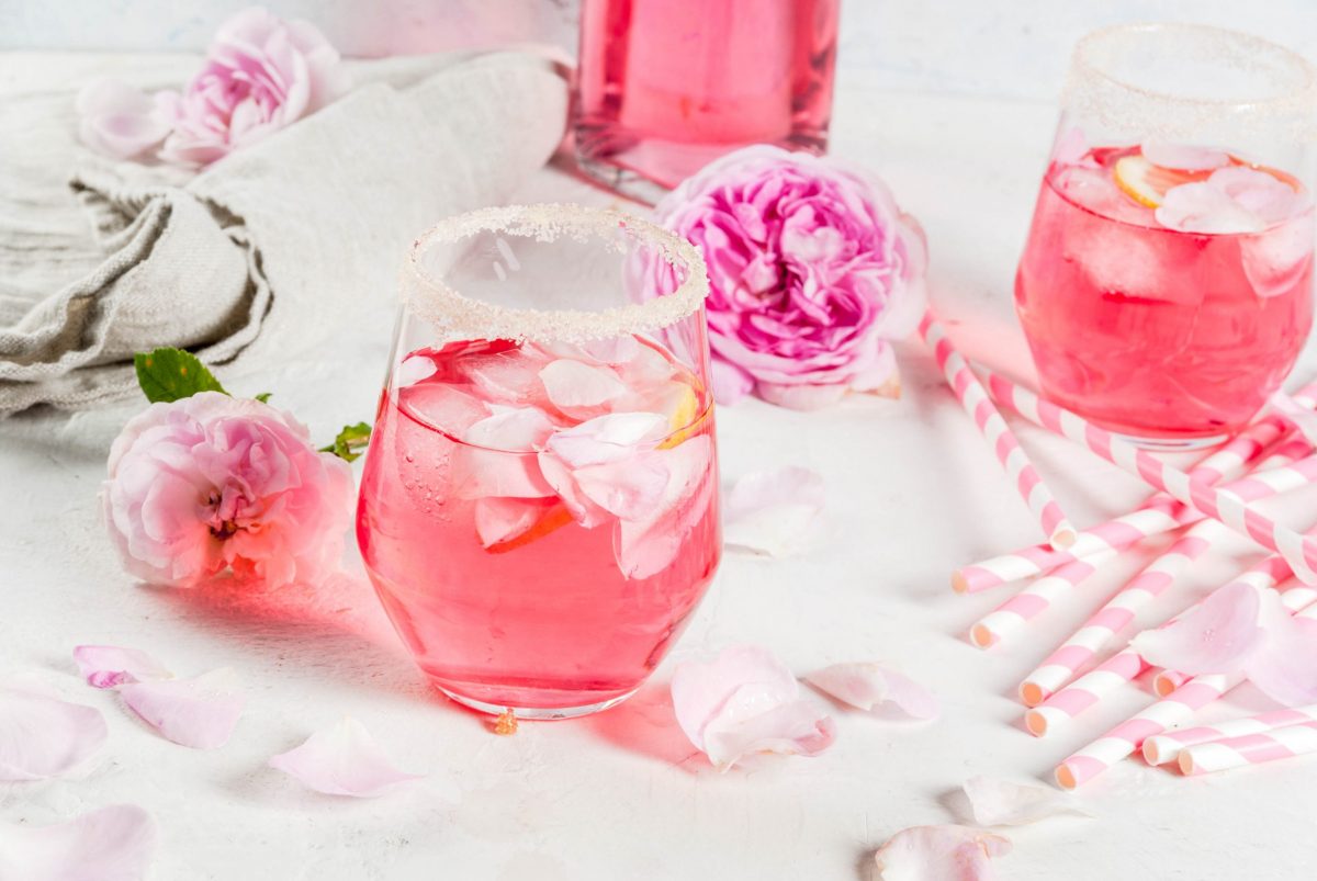 Pink colored cocktail in stem-less wine glass on a white table surrounded by pink flowers and pink striped straws.