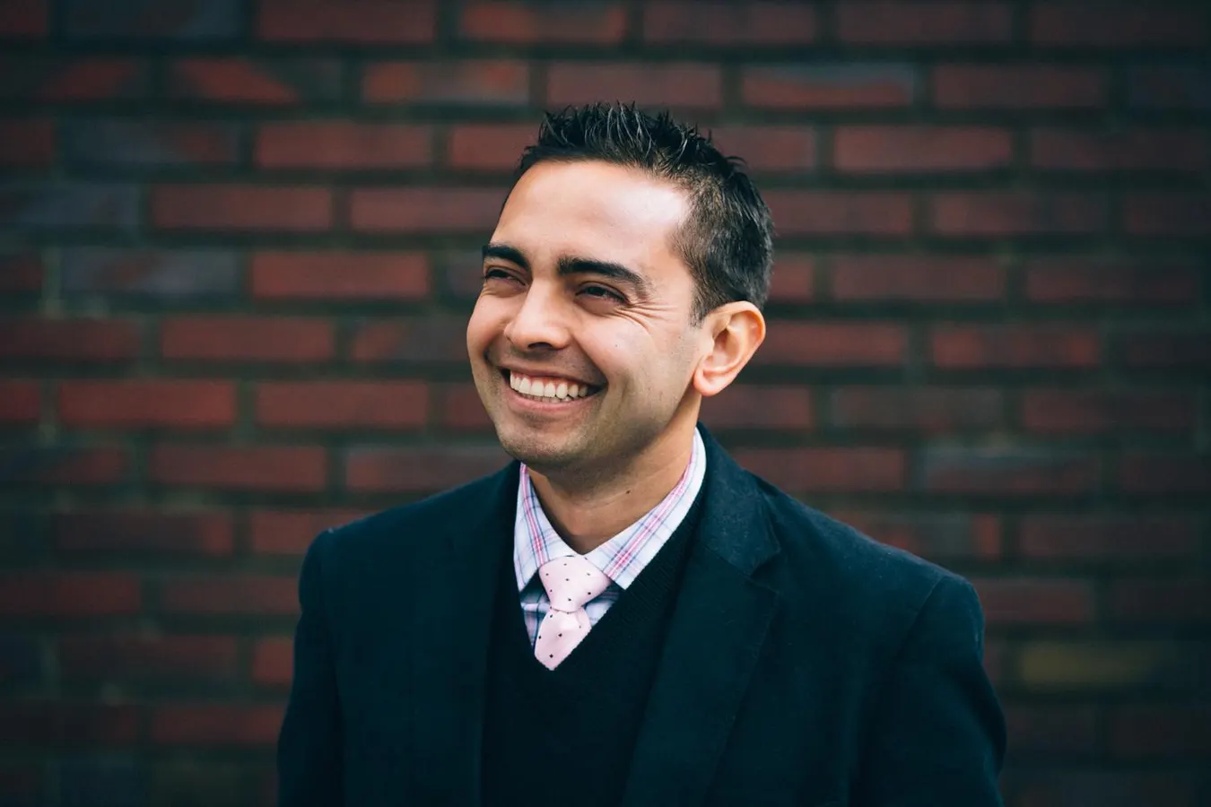 Man with short black hair and medium toned skin smiling and wearing a black suit and white collared shirt standing in front of a dark brick wall.