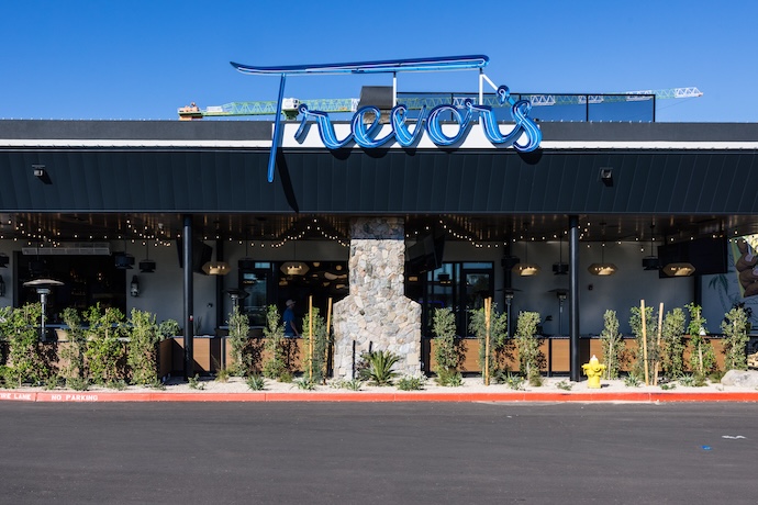 Outside of restaurant store front with black roofing paneling, grey stone structure, and green shrubbery with a blue neon sign on the roof reading "Trevor's"