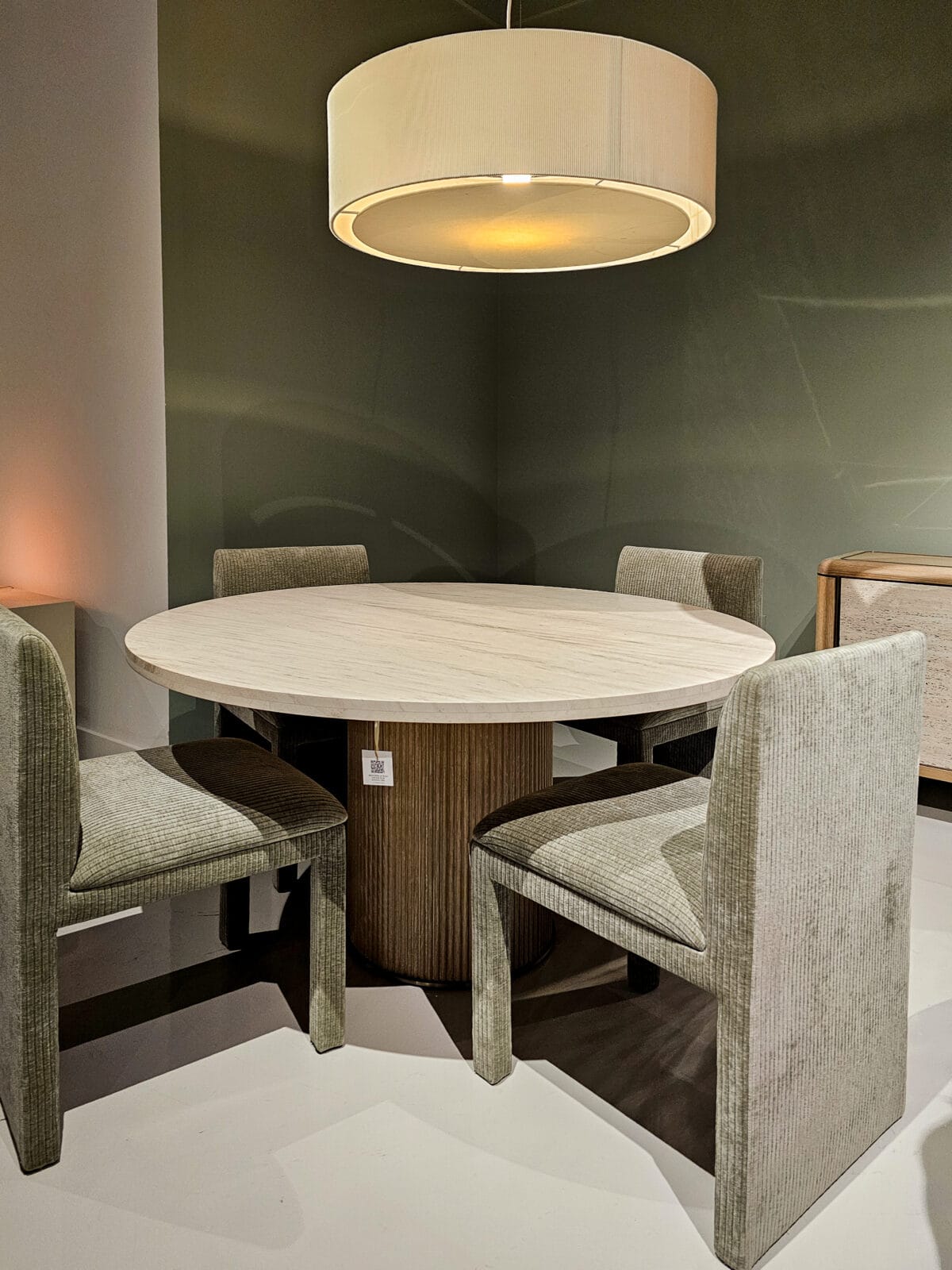 A round wooden dining table with four upholstered chairs is placed under a large circular pendant light in a modern, minimalist room.